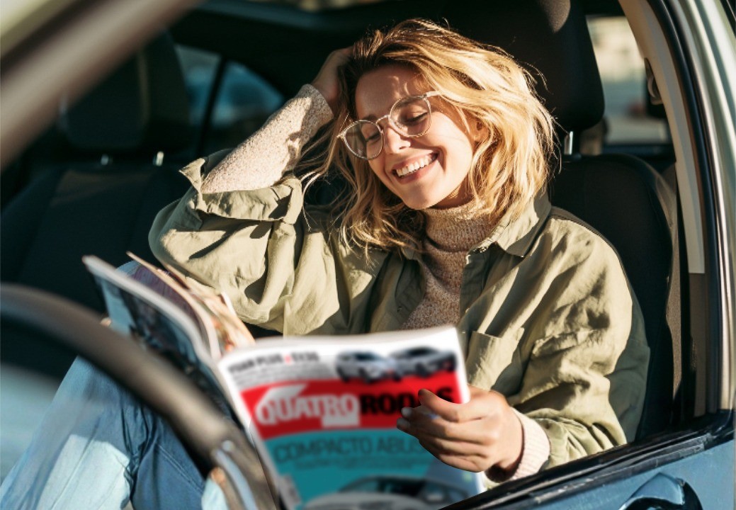Mulher loira, de óculos e blusa verde-oliva, sorri enquanto lê a revista Quatro Rodas dentro de um carro, com a mão direita nos cabelos