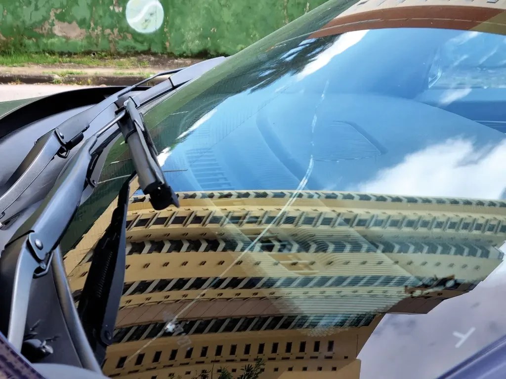 Para-brisa de carro com rachadura vertical, refletindo um prédio bege com janelas escuras e o céu azul com nuvens brancas. Limpadores de para-brisa pretos estão à esquerda, e uma parede verde com grafite branco aparece ao fundo