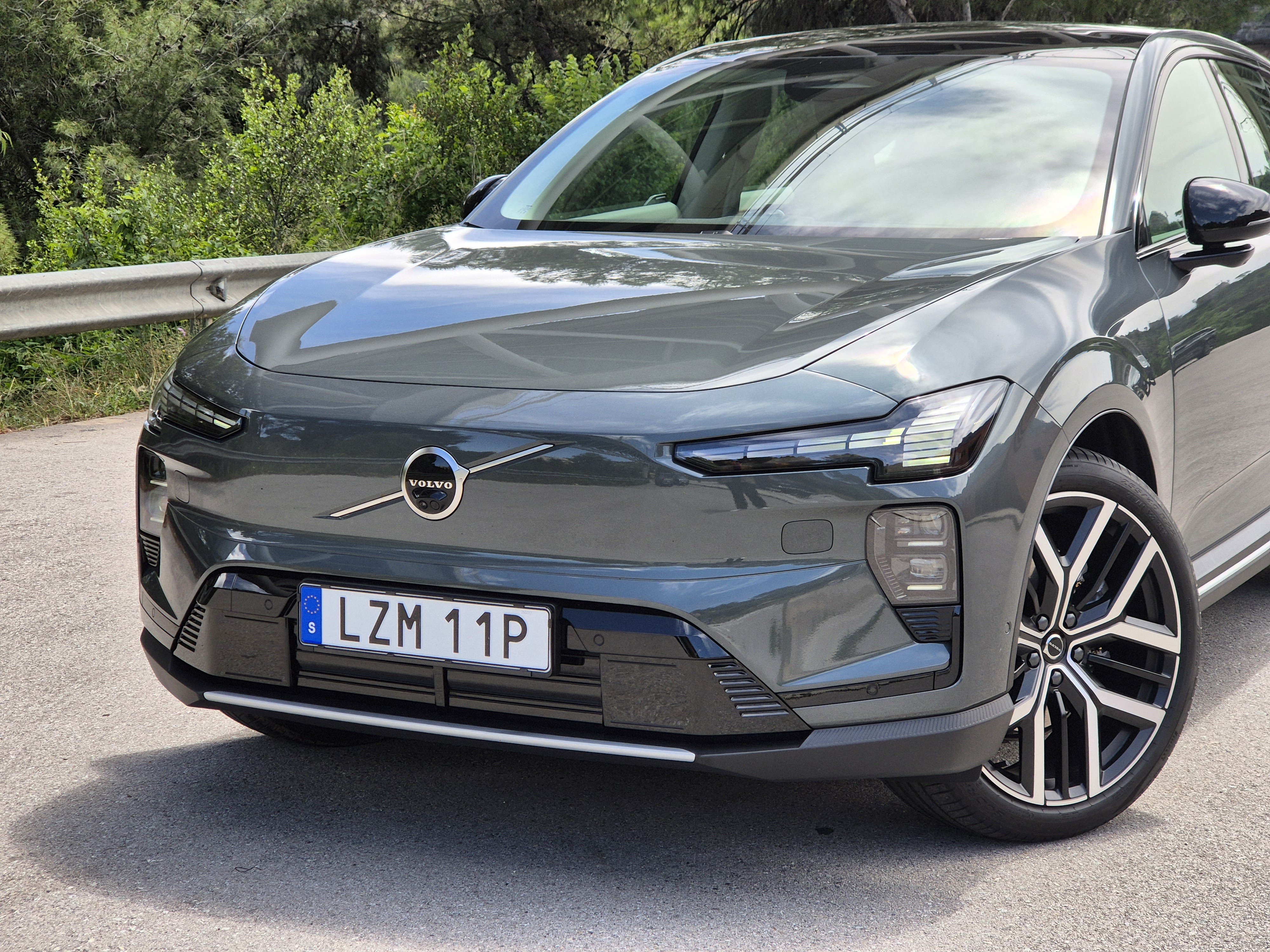 Frente de um Volvo EX60 cinza escuro, com far&oacute;is e luzes diurnas acesas, estacionado em uma estrada asfaltada com vegeta&ccedil;&atilde;o ao fundo