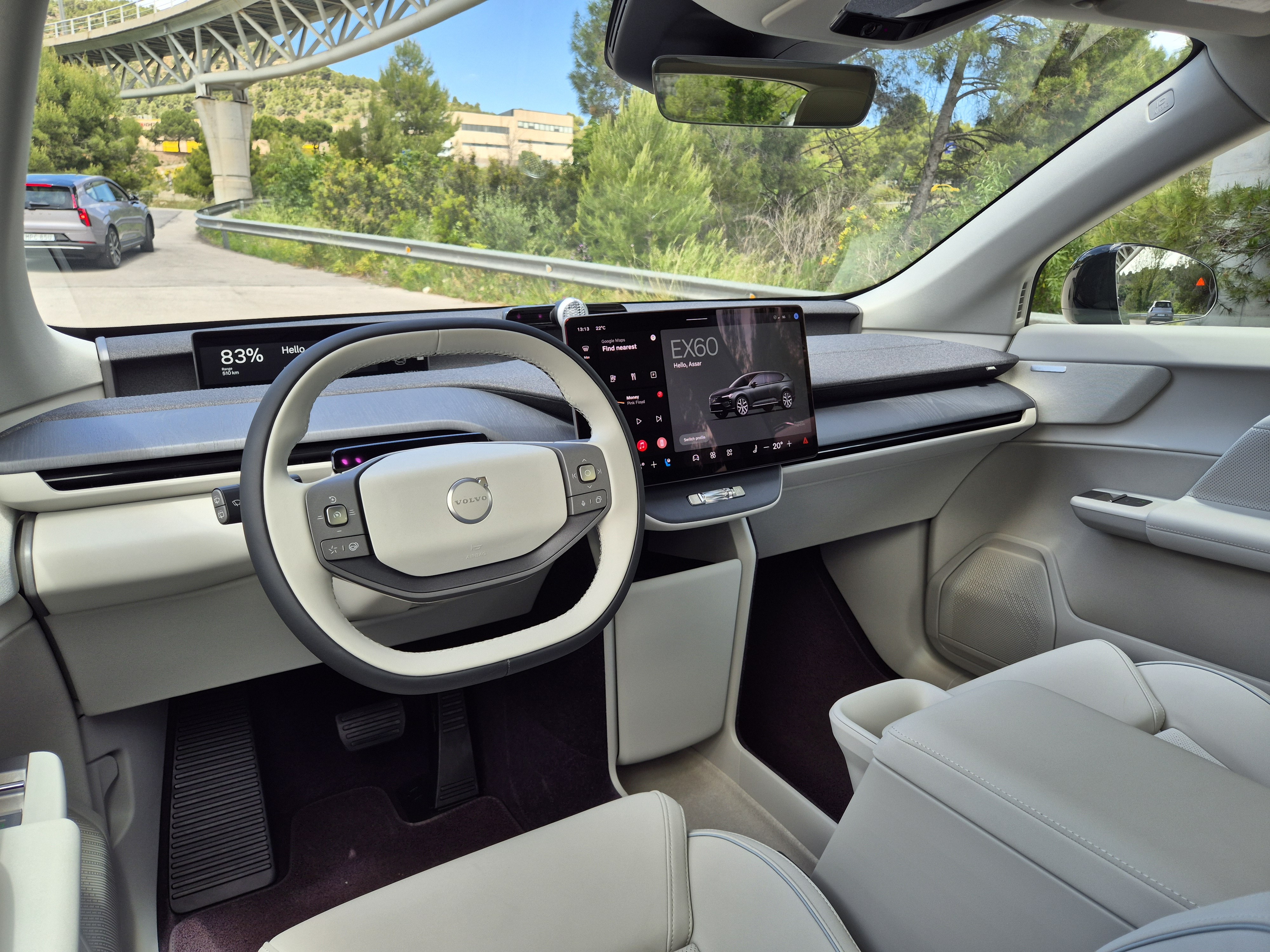 Interior de um Volvo EX60 com volante branco e cinza, painel minimalista e tela central exibindo o modelo. Ao fundo, uma estrada sinuosa com ponte e vegeta&ccedil;&atilde;o verde sob c&eacute;u azul