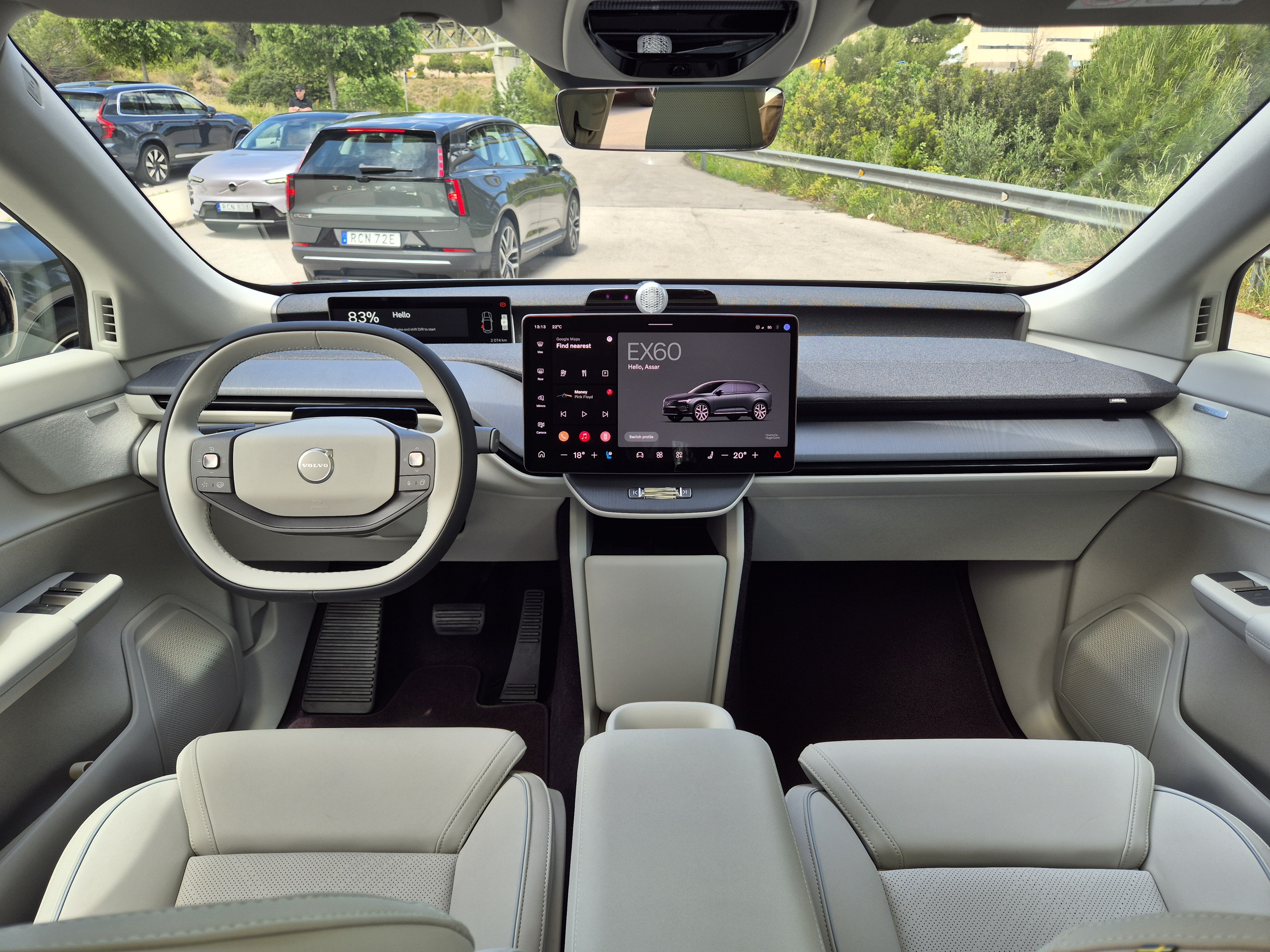 Interior de um carro Volvo EX60, com volante cinza e branco, tela central exibindo o modelo do ve&iacute;culo e carros estacionados ao fundo em uma estrada