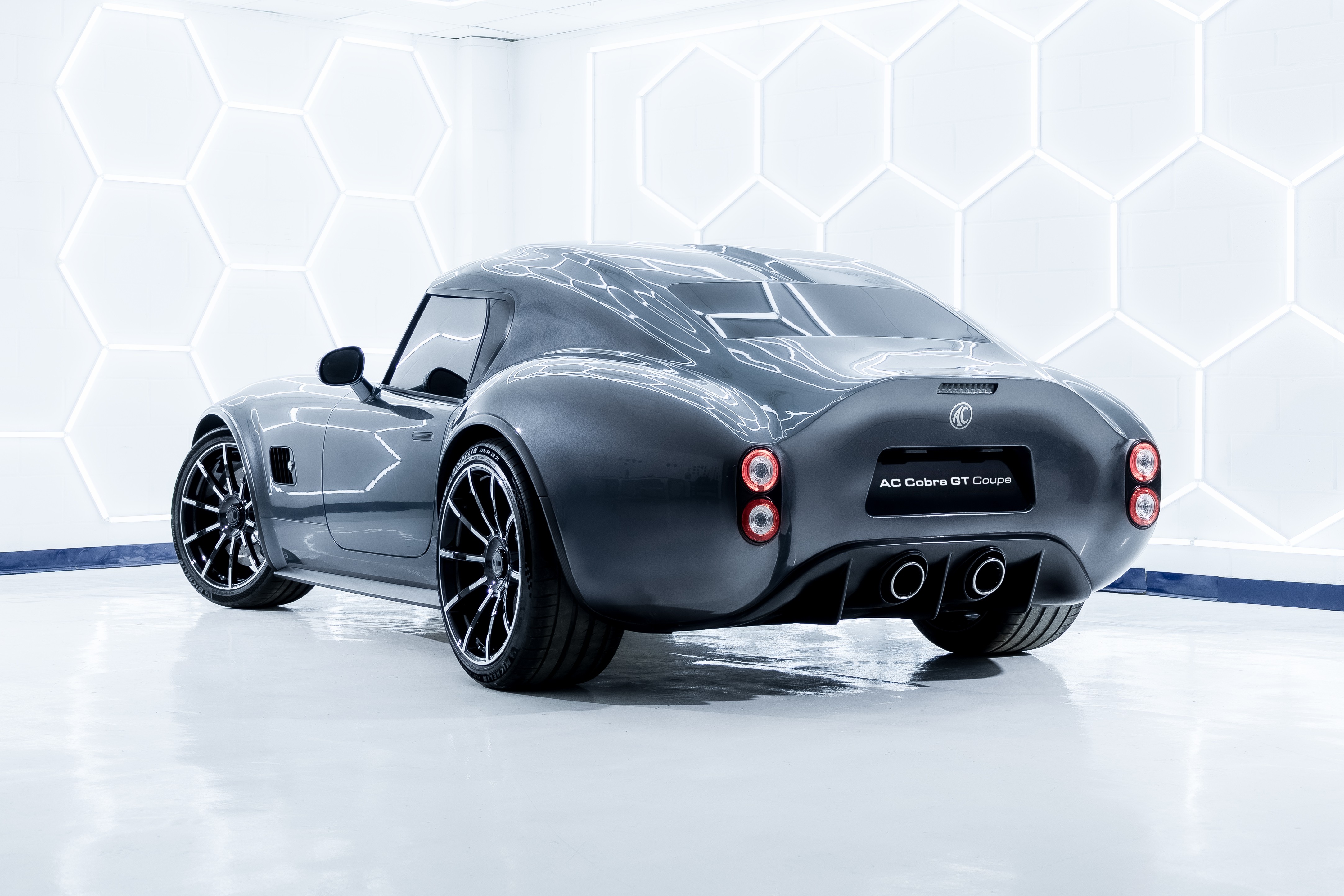 Carro esportivo AC Cobra GT Coupe cinza escuro, visto por tr&aacute;s, com rodas pretas e detalhes cromados, em um est&uacute;dio branco com ilumina&ccedil;&atilde;o hexagonal
