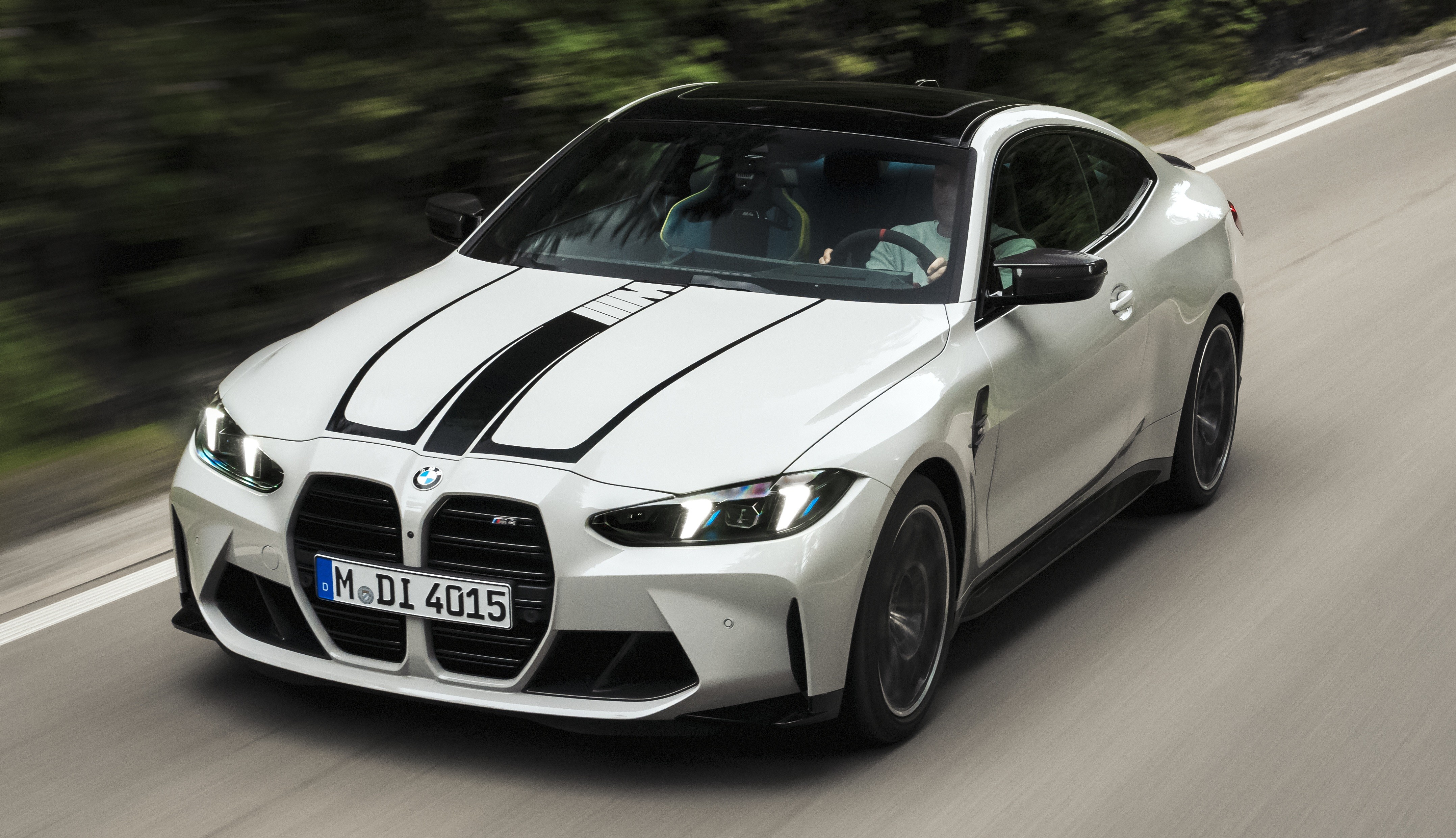 BMW M4 branco com teto preto e duas faixas pretas no cap&ocirc;, em movimento r&aacute;pido numa estrada asfaltada com &aacute;rvores borradas ao fundo
