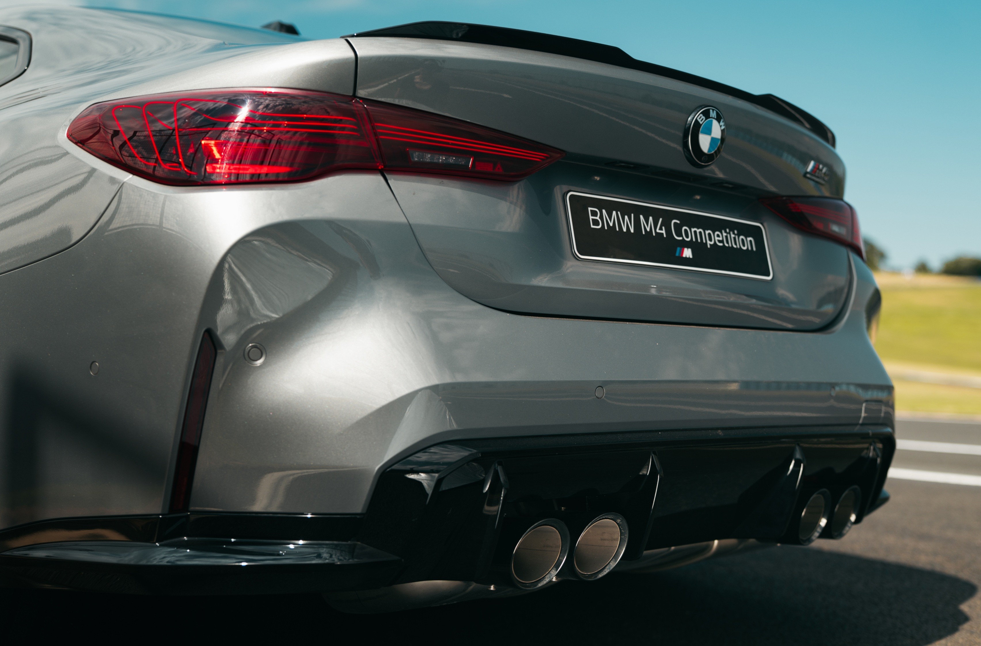 Traseira de um carro BMW M4 Competition cinza met&aacute;lico, com lanternas vermelhas, emblema da marca e quatro sa&iacute;das de escapamento cromadas, em uma pista de corrida sob c&eacute;u azul