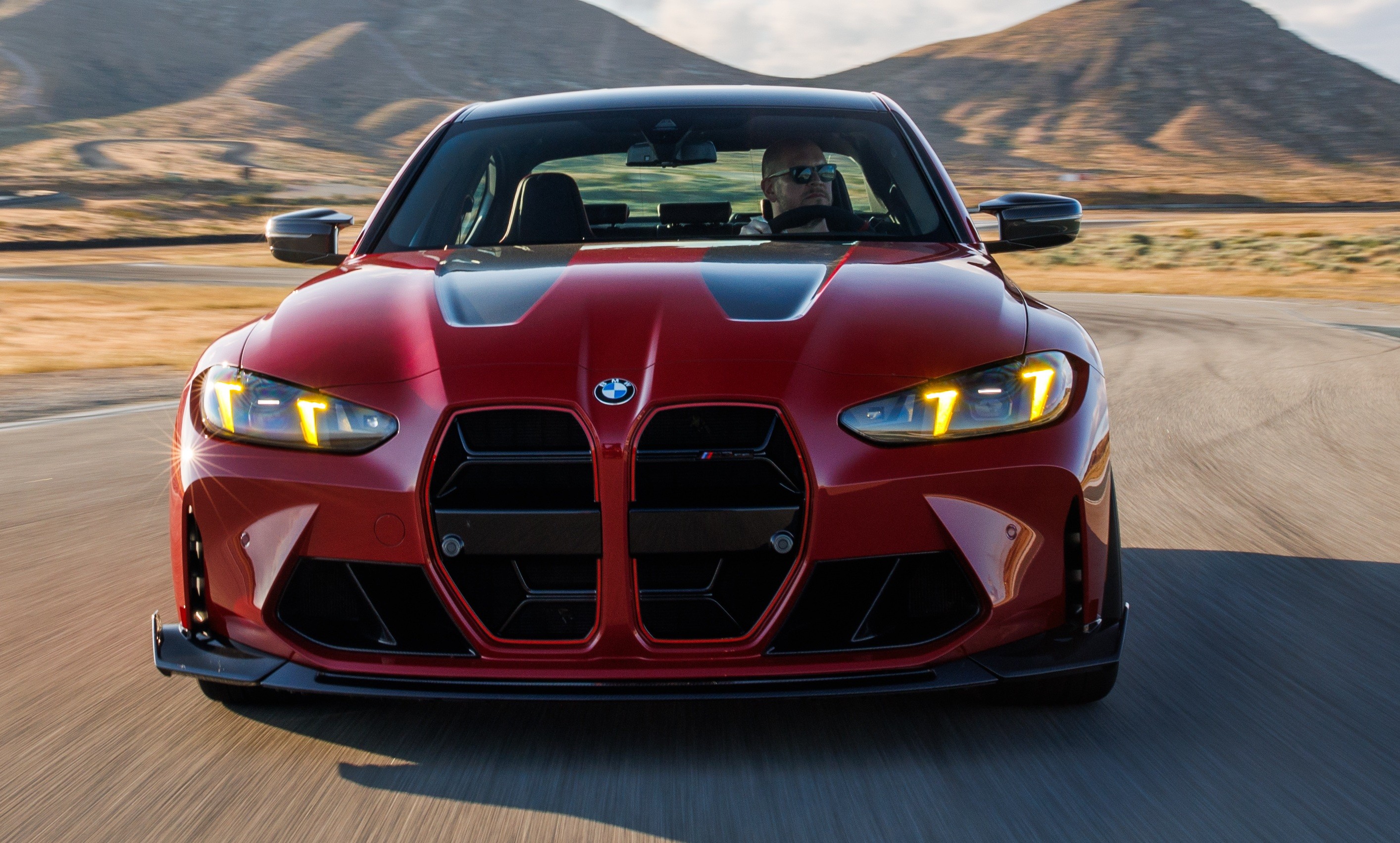 Carro esportivo vermelho BMW M4 Competition, com motorista usando &oacute;culos escuros, em alta velocidade numa pista sinuosa com montanhas ao fundo