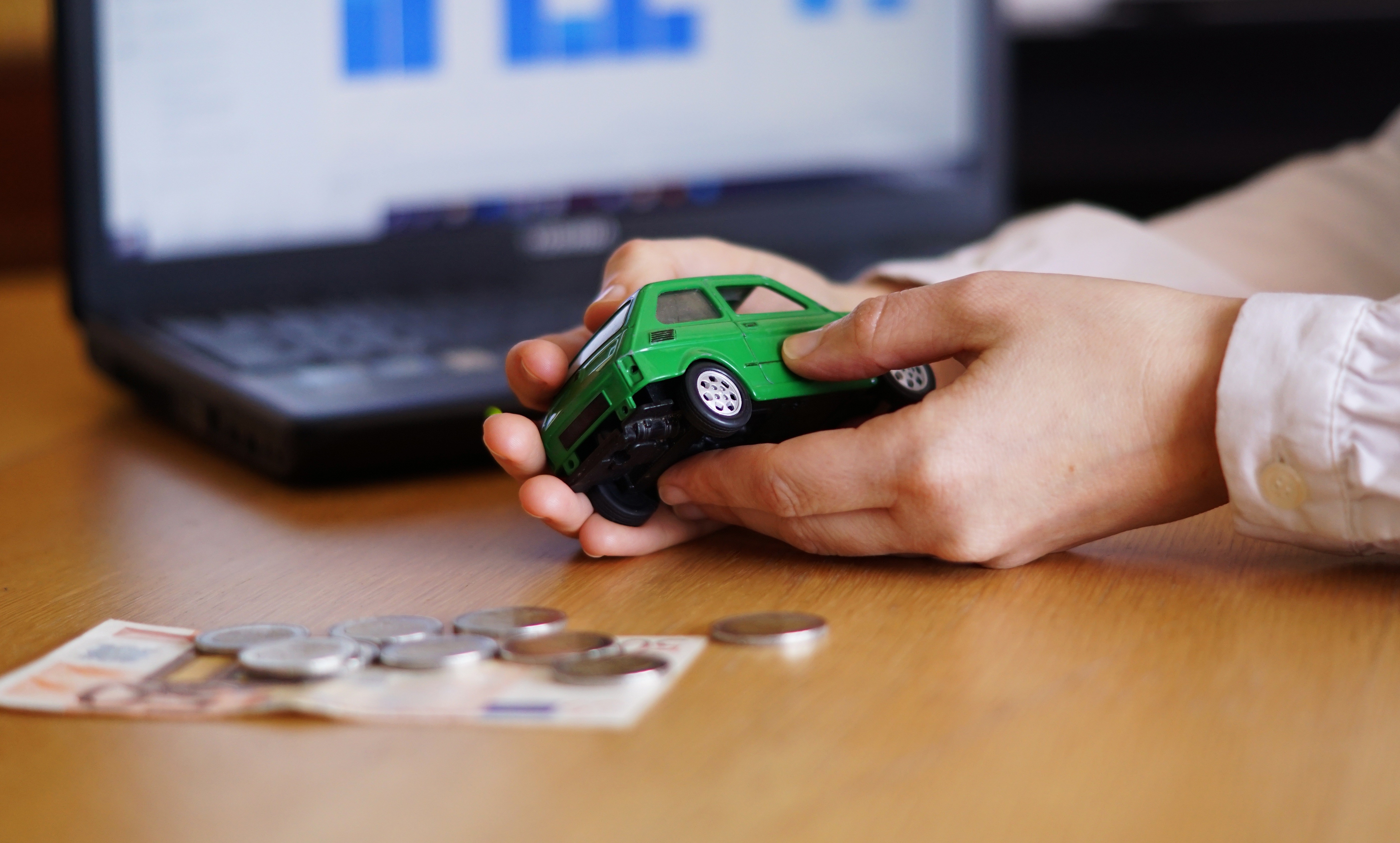 M&atilde;os segurando um carro de brinquedo verde sobre uma mesa de madeira, com notas de euro e moedas espalhadas, e um notebook com gr&aacute;ficos ao fundo