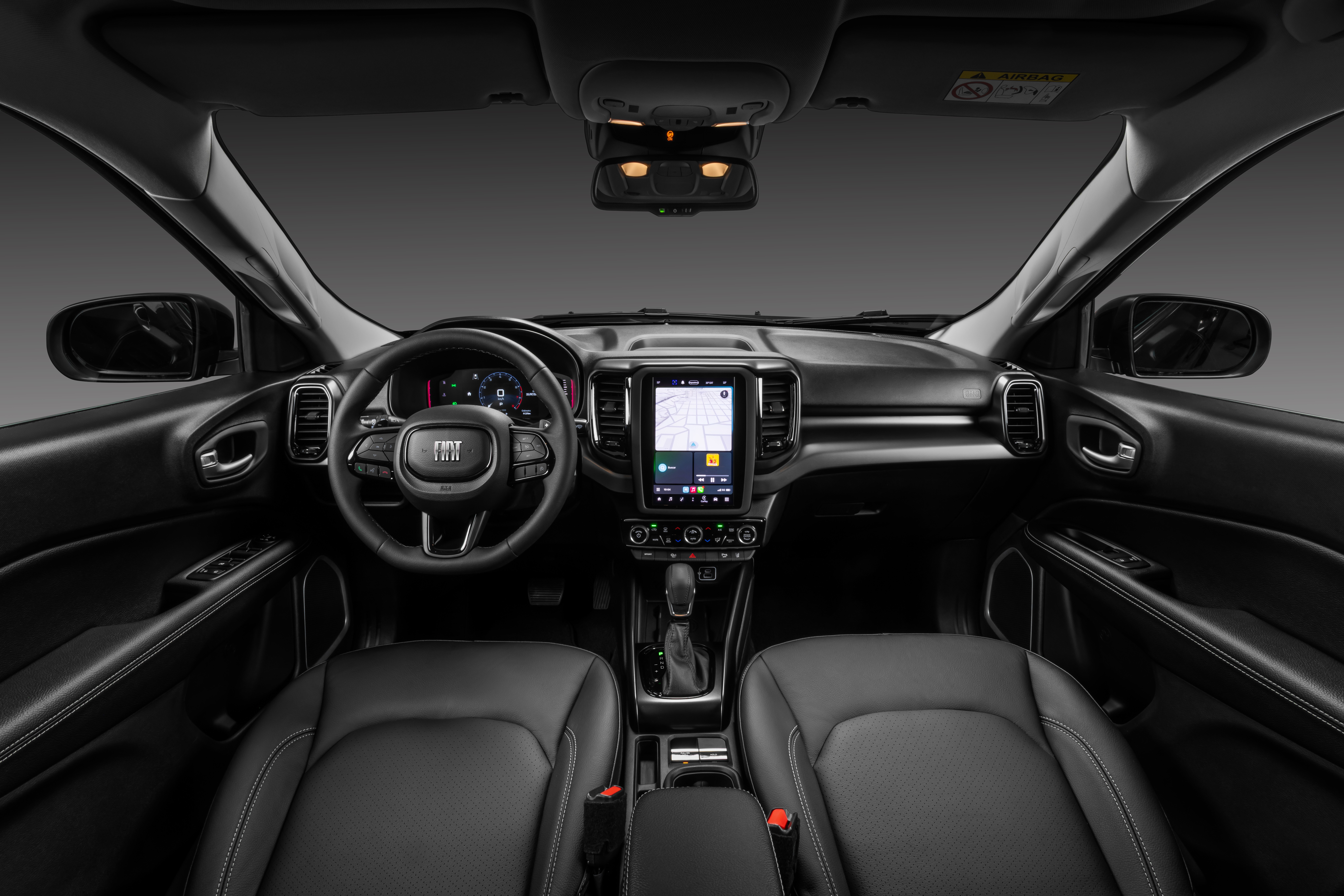 Interior de um carro moderno, com bancos de couro pretos, volante multifuncional com logo Fiat, painel digital e uma tela multim&iacute;dia vertical exibindo um mapa de navega&ccedil;&atilde;o. O c&acirc;mbio autom&aacute;tico est&aacute; no console central, e o espelho retrovisor interno e as luzes de teto s&atilde;o vis&iacute;veis.