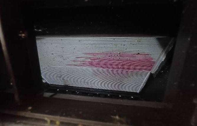 Radiador de carro com aletas met&aacute;licas horizontais e mancha rosa avermelhada de vazamento de fluido, em um compartimento escuro