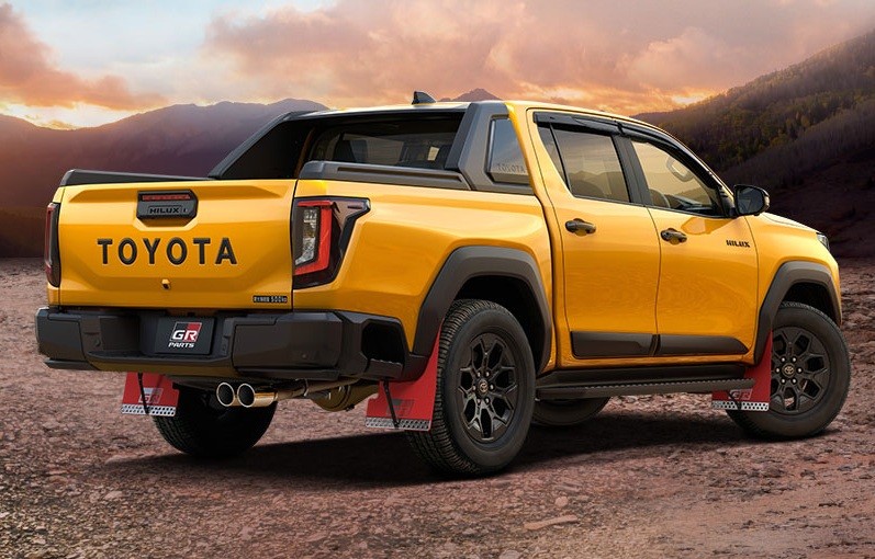 Picape Toyota Hilux amarela, vista de tr&aacute;s e lateral, com detalhes em preto fosco, para-lamas vermelhos e rodas escuras, estacionada em terreno de terra com montanhas ao fundo e c&eacute;u alaranjado ao p&ocirc;r do sol