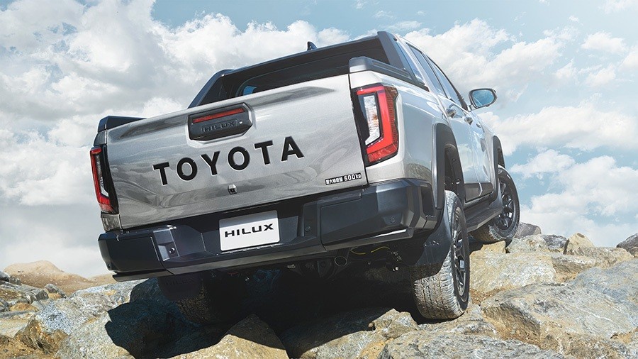 Picape Toyota Hilux prateada vista por tr&aacute;s, subindo uma trilha rochosa sob um c&eacute;u azul com nuvens esparsas