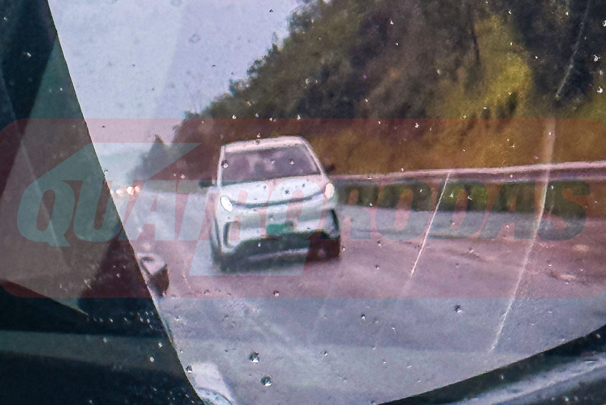 Espelho retrovisor lateral de um carro, molhado pela chuva, refletindo um ve&iacute;culo branco na estrada sob um c&eacute;u nublado e montanhas ao fundo