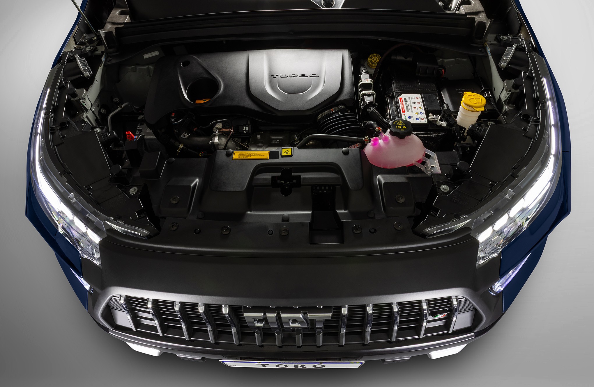 Vista superior do motor de um carro azul escuro com o cap&ocirc; aberto, mostrando o motor turbo, bateria, reservat&oacute;rios de fluidos rosa e amarelo, e a grade frontal com o logo Fiat e a placa 