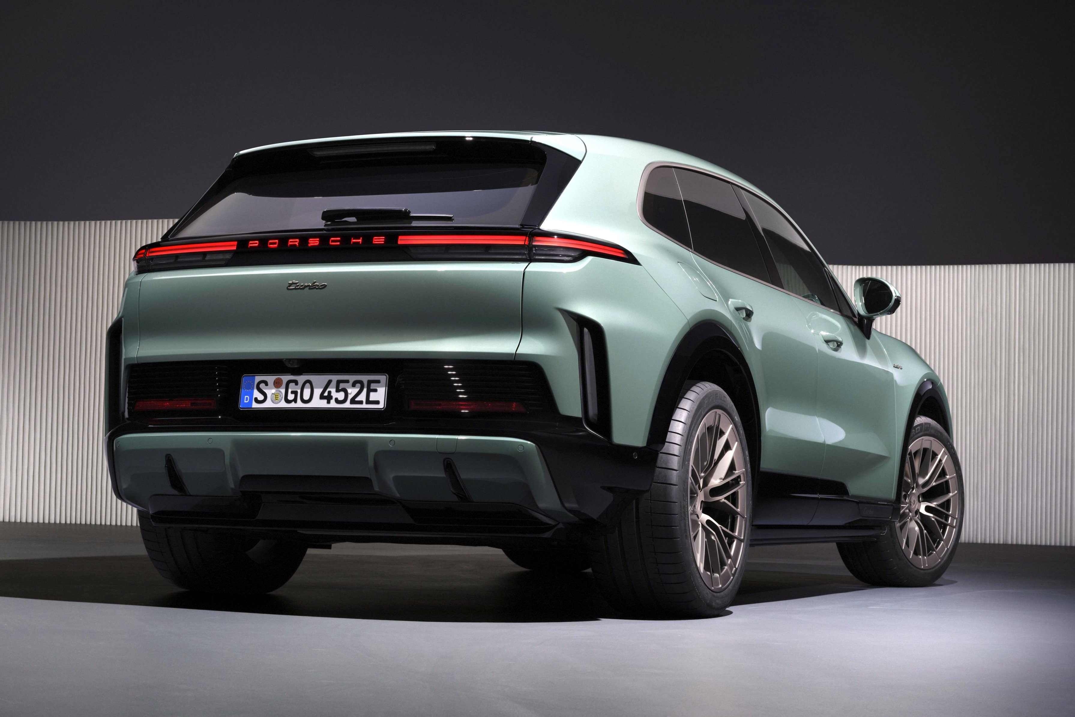 SUV Porsche Cayenne Turbo E-Hybrid verde-claro, visto de traseira e lateral, com lanternas conectadas e placa alem&atilde; S GO 452E, em ambiente escuro com parede listrada
