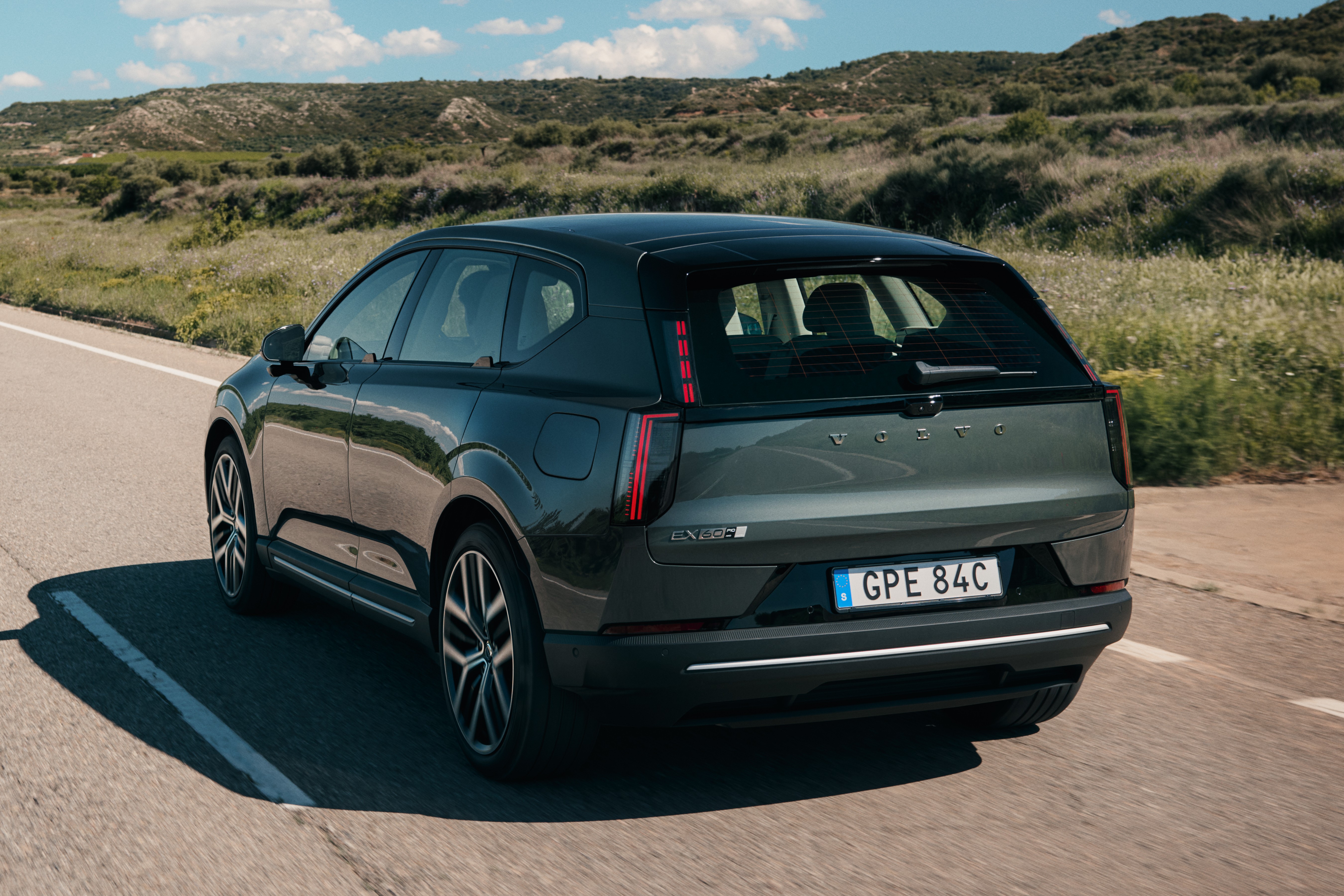 SUV Volvo EX30 cinza-escuro, visto de tr&aacute;s, em uma estrada asfaltada com faixa branca. Ao fundo, colinas verdes e c&eacute;u azul com nuvens esparsas