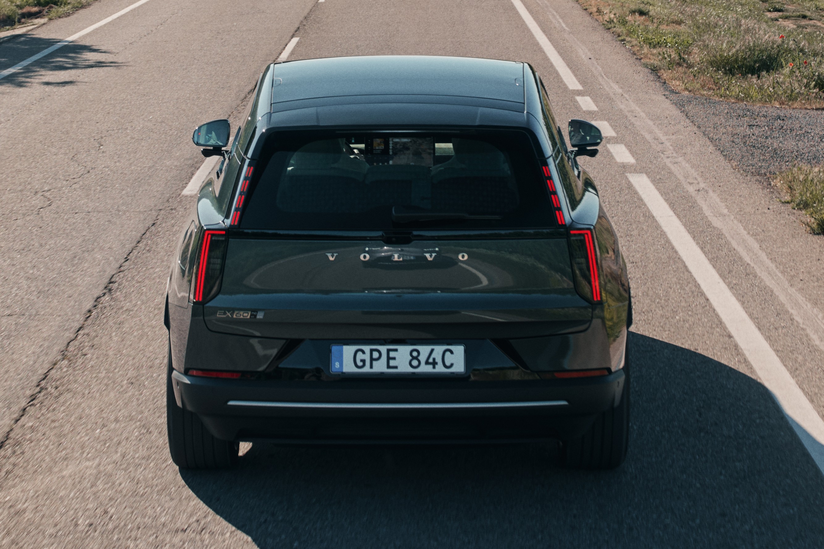 Traseira de um carro Volvo preto em uma estrada asfaltada com faixas brancas, sob luz solar, com vegeta&ccedil;&atilde;o seca &agrave; direita