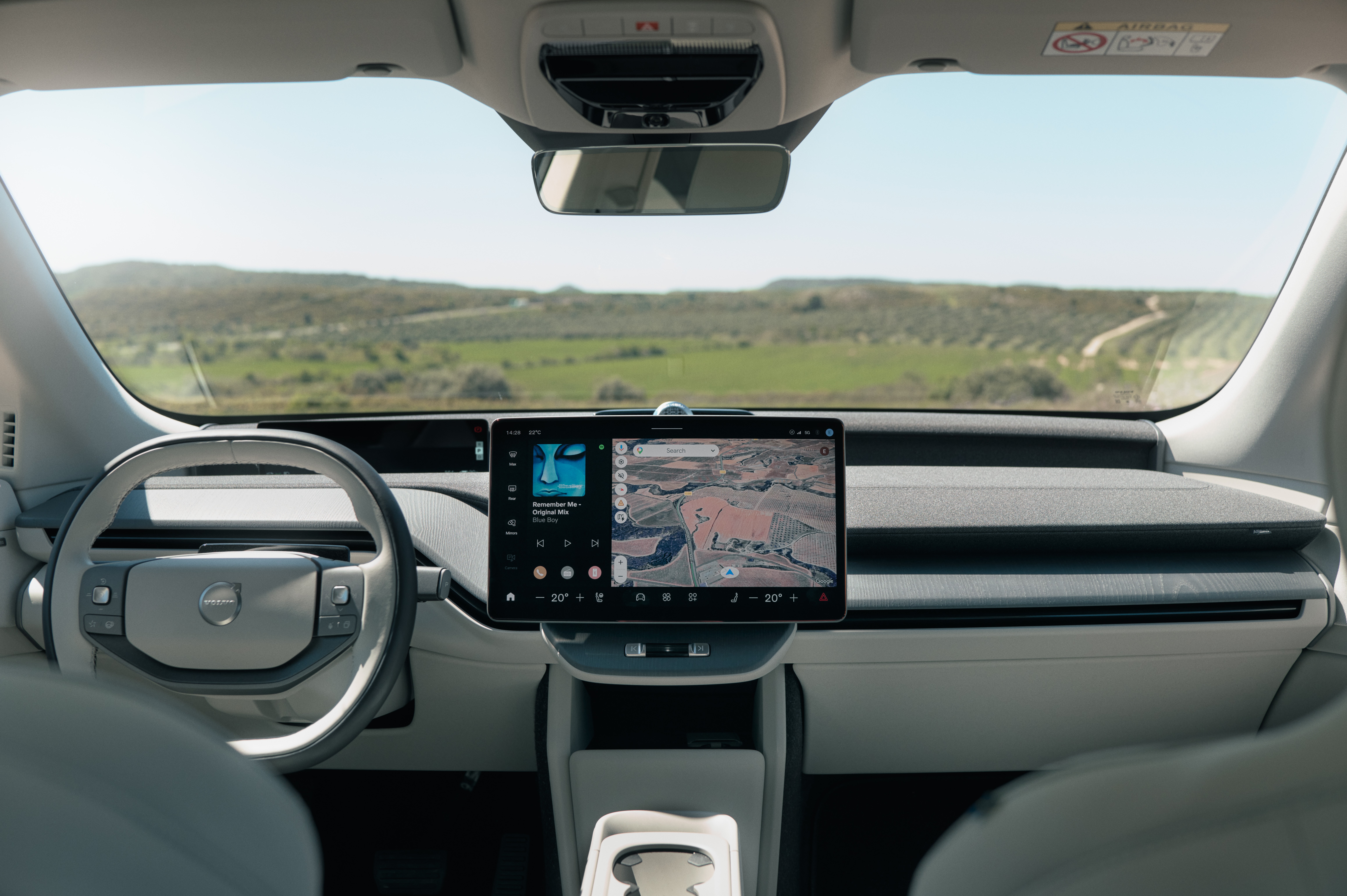 Interior de um carro moderno com volante cinza claro &agrave; esquerda e uma tela central exibindo um mapa e reprodutor de m&uacute;sica. O para-brisa revela uma paisagem verde de colinas sob c&eacute;u azul