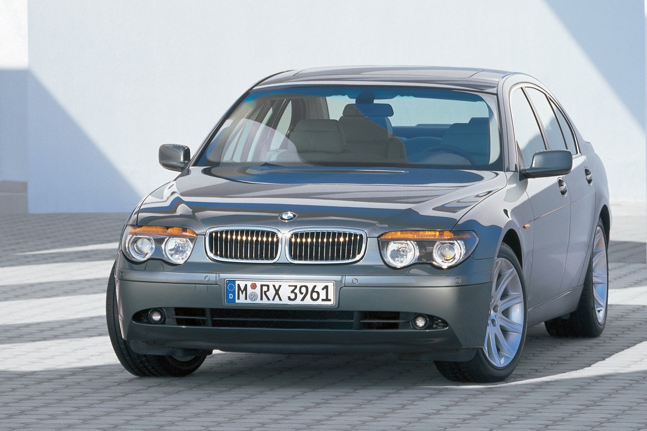 Carro BMW S&eacute;rie 7 cinza escuro estacionado em uma superf&iacute;cie de blocos, com uma parede branca ao fundo e sombra projetada