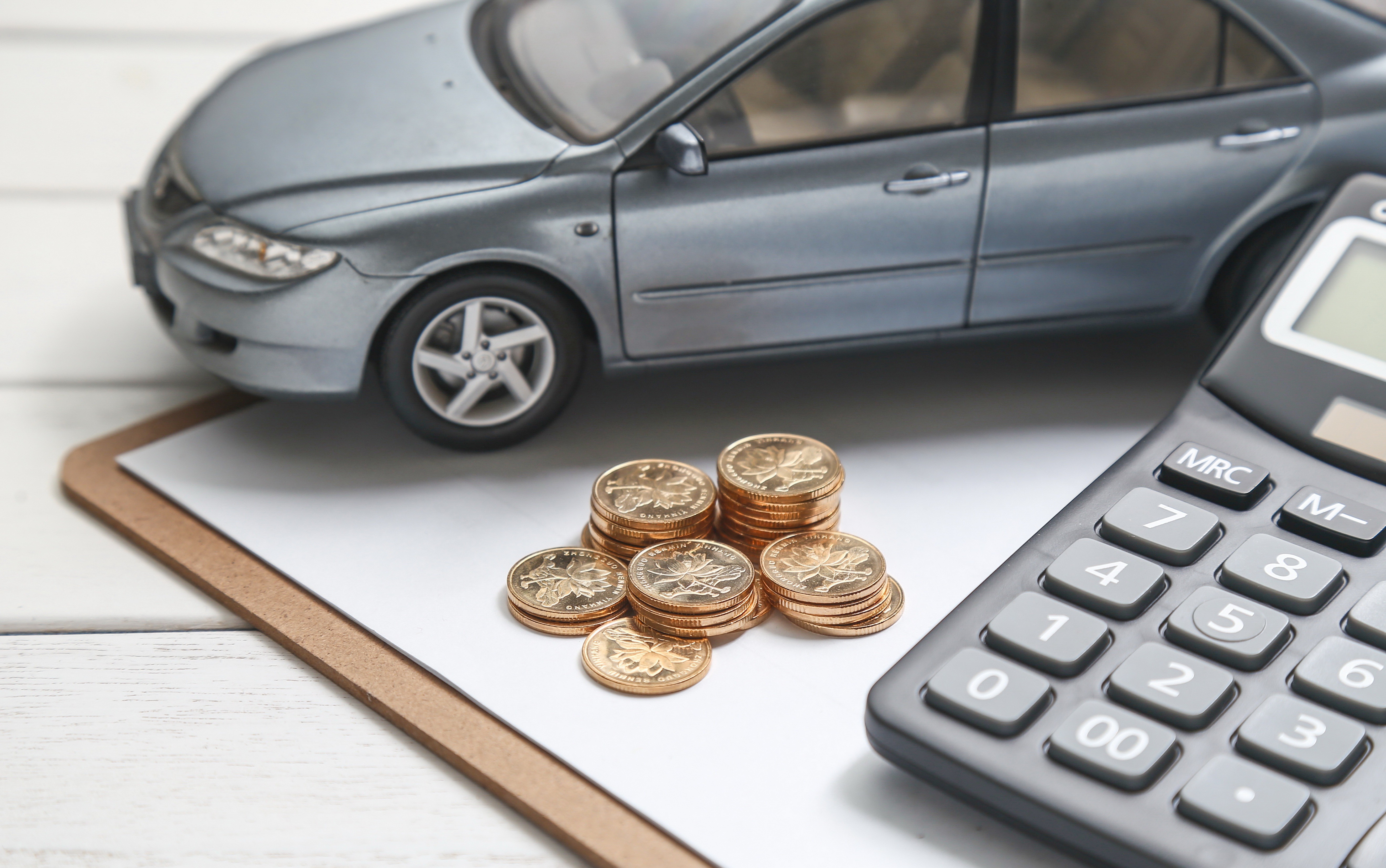 Miniatura de carro cinza sobre prancheta branca, com pilhas de moedas douradas e uma calculadora preta &agrave; direita, simbolizando custos veiculares