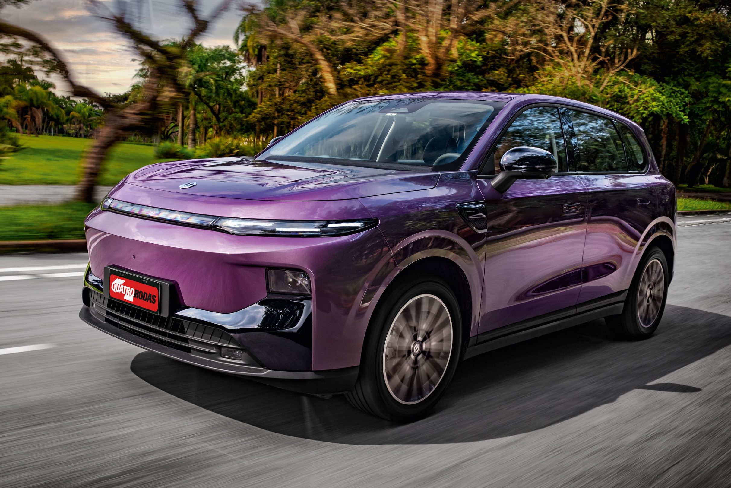 SUV elétrico roxo escuro em movimento, visto de frente e lateral, com rodas girando e fundo de árvores borrado, indicando velocidade