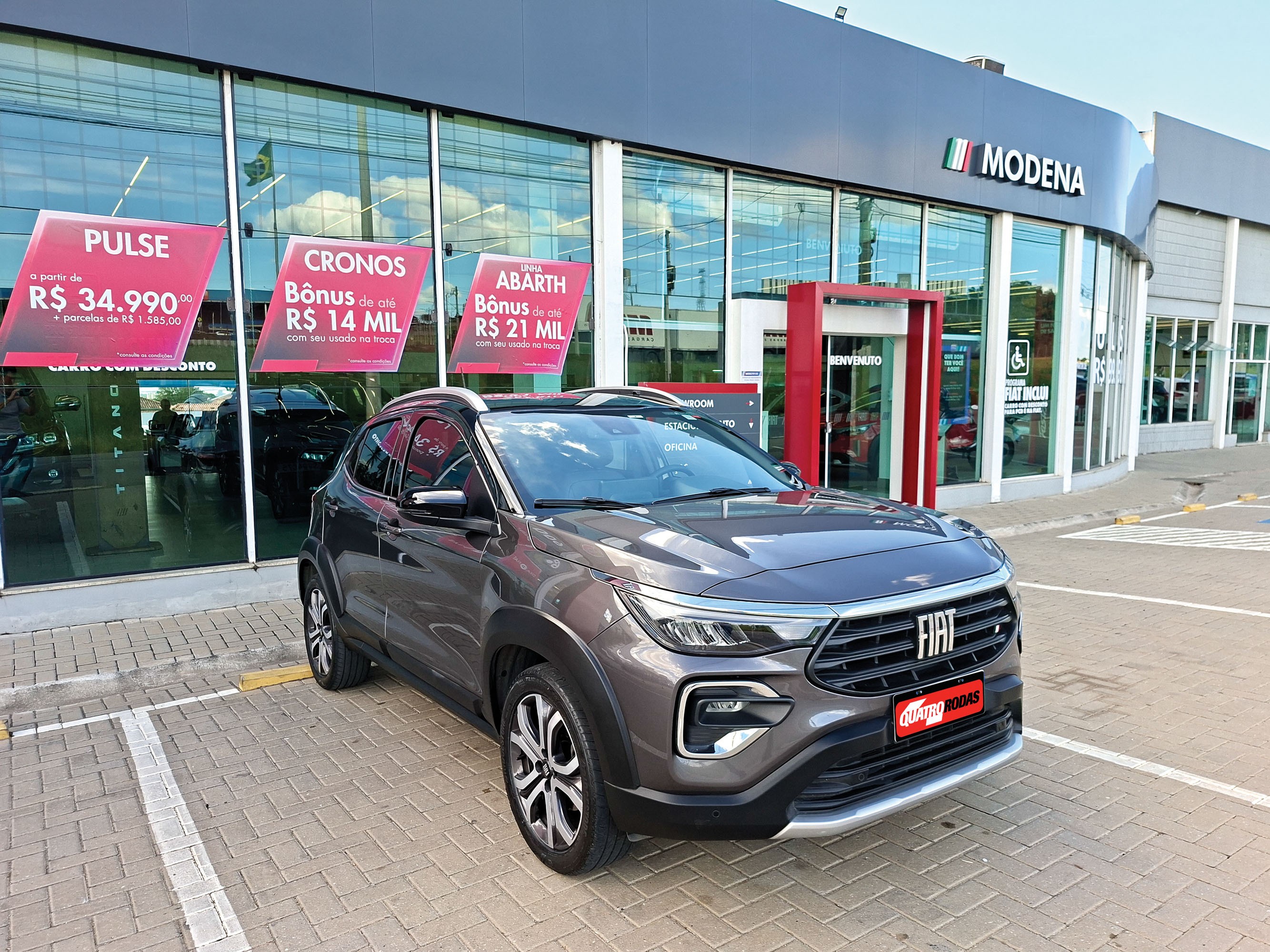 Carro Fiat Pulse cinza-escuro estacionado em frente a uma concessionária Modena com fachada de vidro, exibindo banners de ofertas para modelos Pulse, Cronos e Abarth
