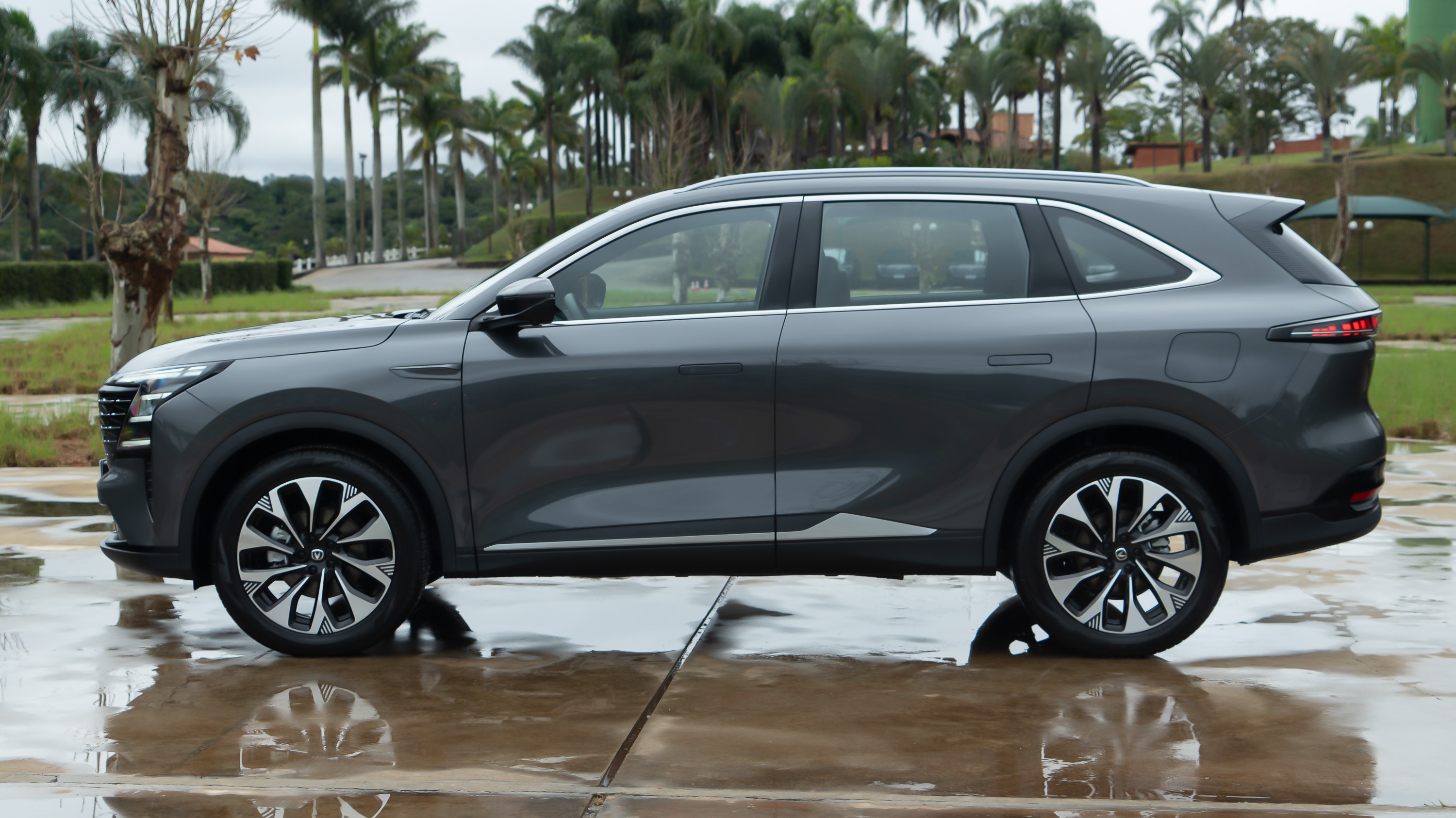 SUV cinza escuro estacionado em uma superf&iacute;cie molhada, com reflexos da &aacute;gua. Ao fundo, palmeiras e vegeta&ccedil;&atilde;o verde sob um c&eacute;u nublado