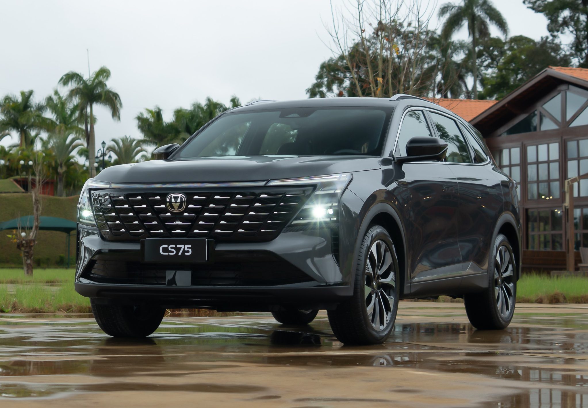 SUV Changan CS75 Plus cinza-escuro, com far&oacute;is acesos, estacionado em p&aacute;tio molhado. Ao fundo, gramado, palmeiras, uma torre verde e uma casa de madeira com telhado de cer&acirc;mica