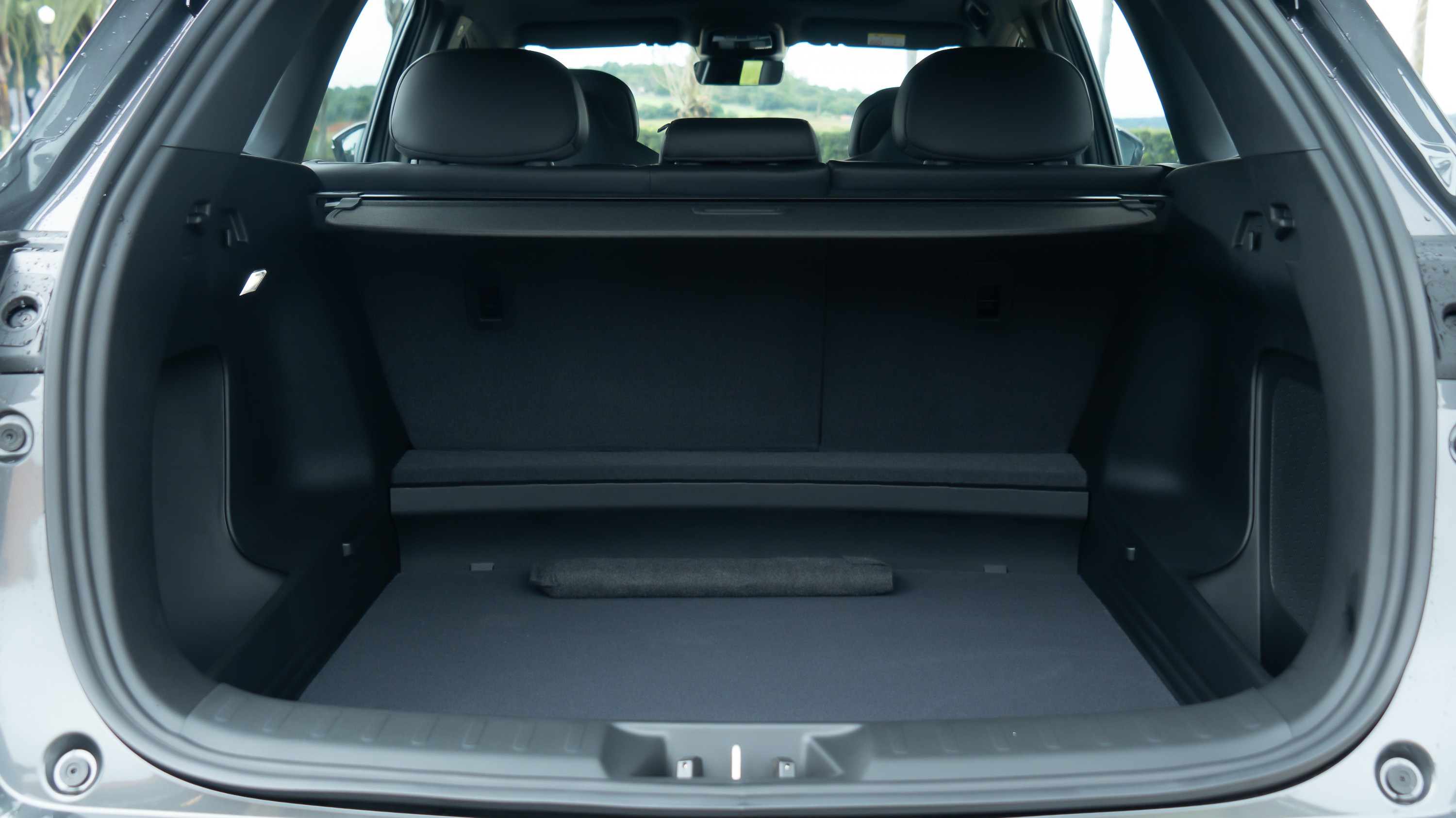 Porta-malas de um carro cinza escuro, visto de tr&aacute;s, com o compartimento vazio e o banco traseiro rebatido. O interior &eacute; preto, com uma cobertura retr&aacute;til sobre o espa&ccedil;o do porta-malas. Ao fundo, os encostos de cabe&ccedil;a dos bancos dianteiros s&atilde;o vis&iacute;veis, e atrav&eacute;s do para-brisa traseiro, um cen&aacute;rio desfocado de &aacute;rvores e c&eacute;u.