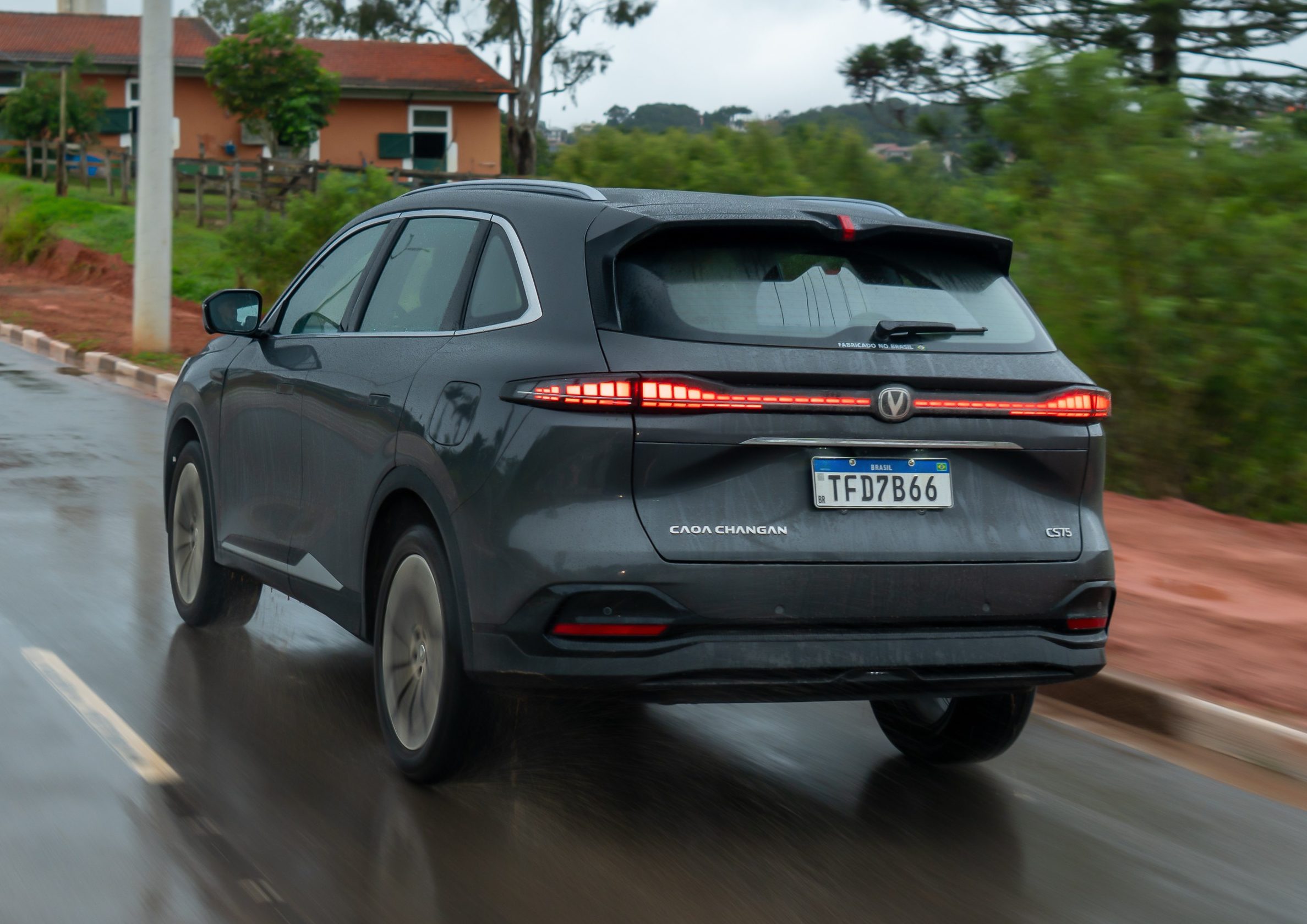 SUV cinza escuro, com lanternas traseiras acesas em LED, trafegando em uma estrada molhada. Ao fundo, casas e vegeta&ccedil;&atilde;o verde sob c&eacute;u nublado