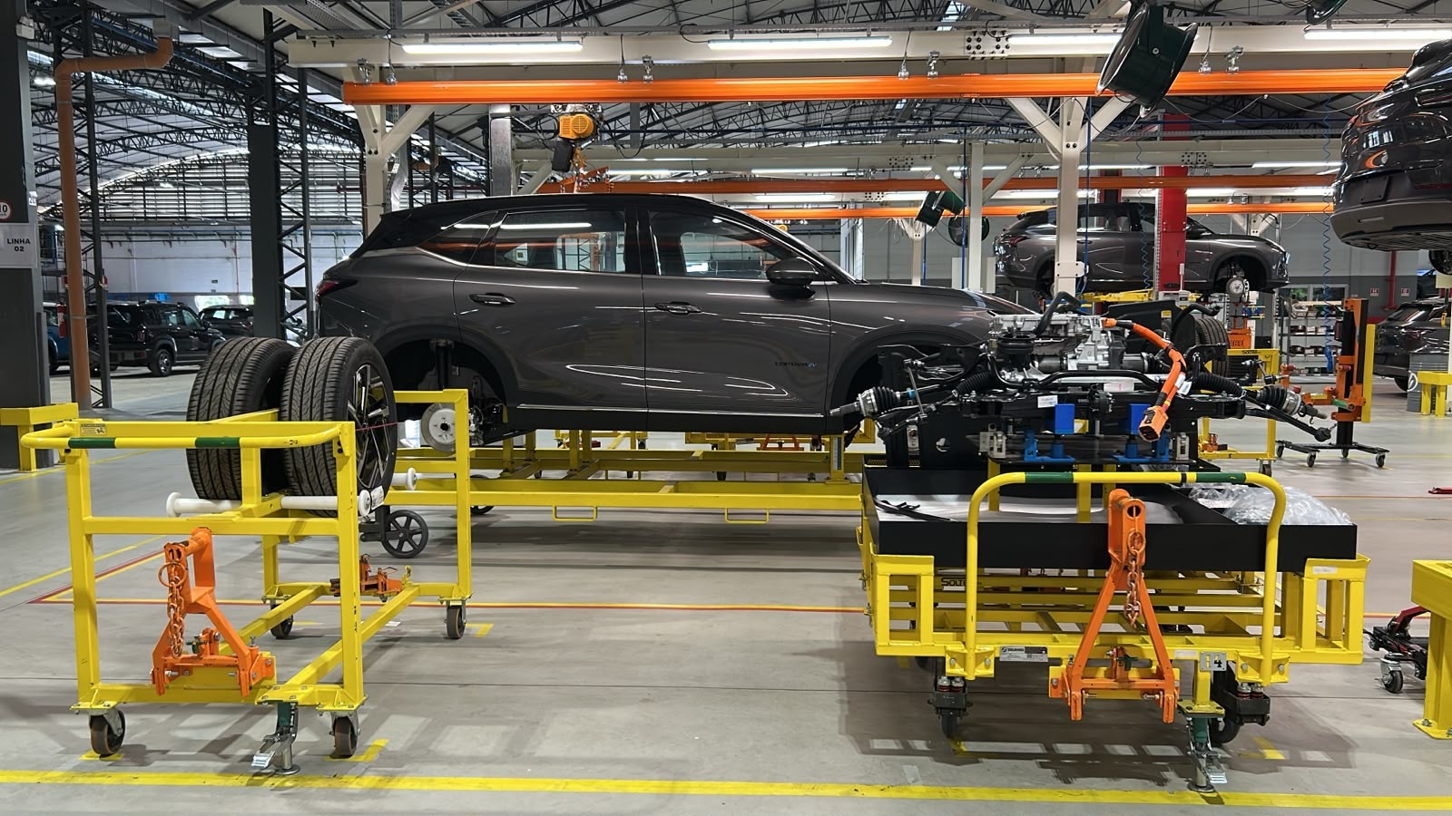 Linha de montagem de ve&iacute;culos com um carro cinza escuro suspenso em uma plataforma amarela, ao lado de um chassi com motor e componentes el&eacute;tricos, tamb&eacute;m em plataforma amarela. Pneus novos est&atilde;o em um carrinho amarelo &agrave; esquerda, e outros carros s&atilde;o vis&iacute;veis ao fundo, em diferentes est&aacute;gios de montagem
