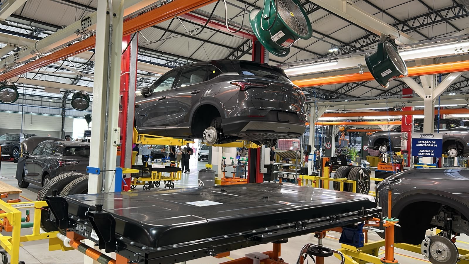 Linha de montagem de ve&iacute;culos el&eacute;tricos, com um carro cinza suspenso sem rodas e uma grande bateria preta no primeiro plano, pronta para instala&ccedil;&atilde;o. Outros carros em diferentes est&aacute;gios de montagem e trabalhadores ao fundo.