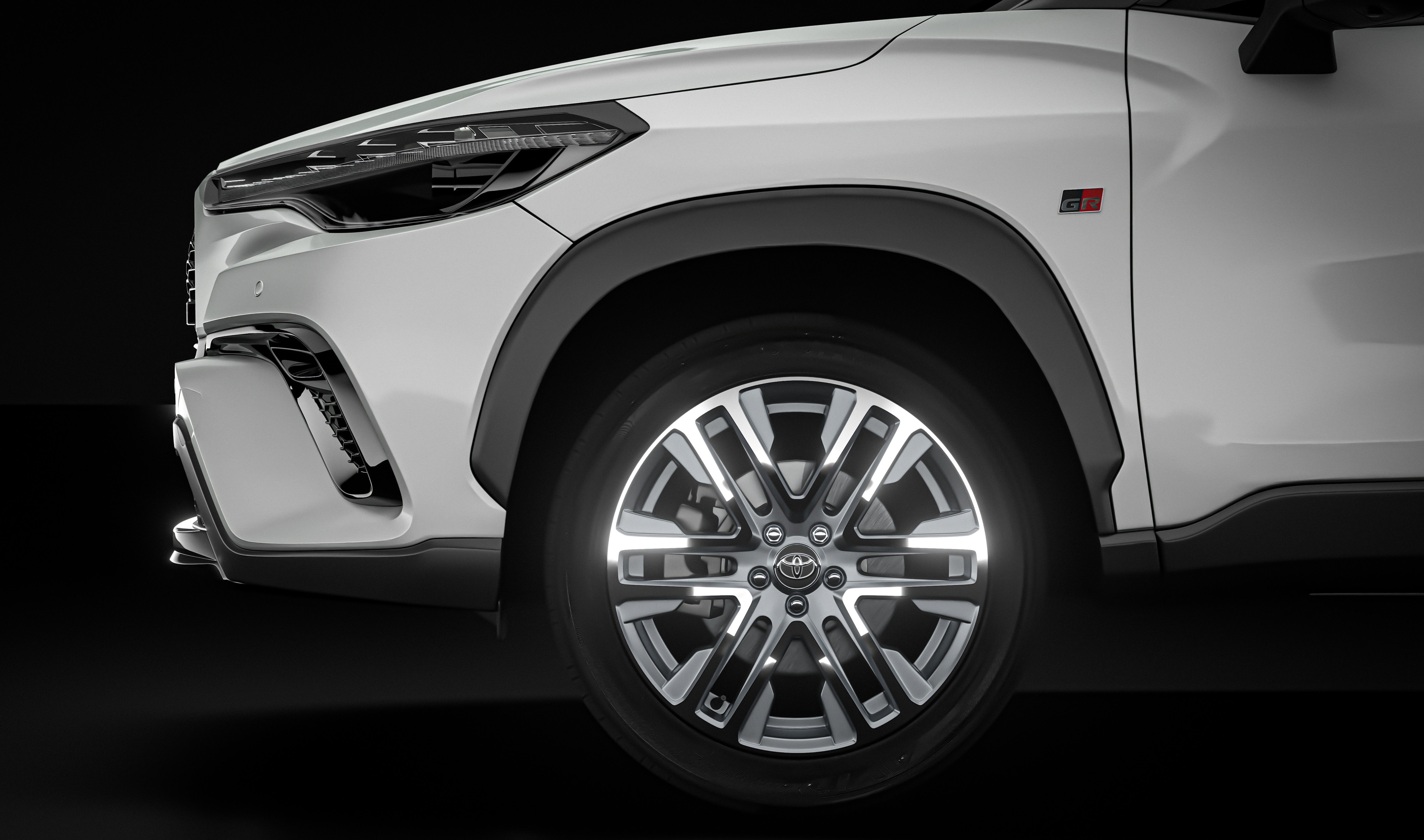 Lateral dianteira de um carro branco com detalhes pretos, incluindo o para-choque, a caixa de roda e a saia. A roda possui aros prateados e pretos, com o emblema da Toyota no centro. Um pequeno distintivo GR vermelho e preto est&aacute; vis&iacute;vel acima da roda, no para-lama. O fundo &eacute; totalmente preto