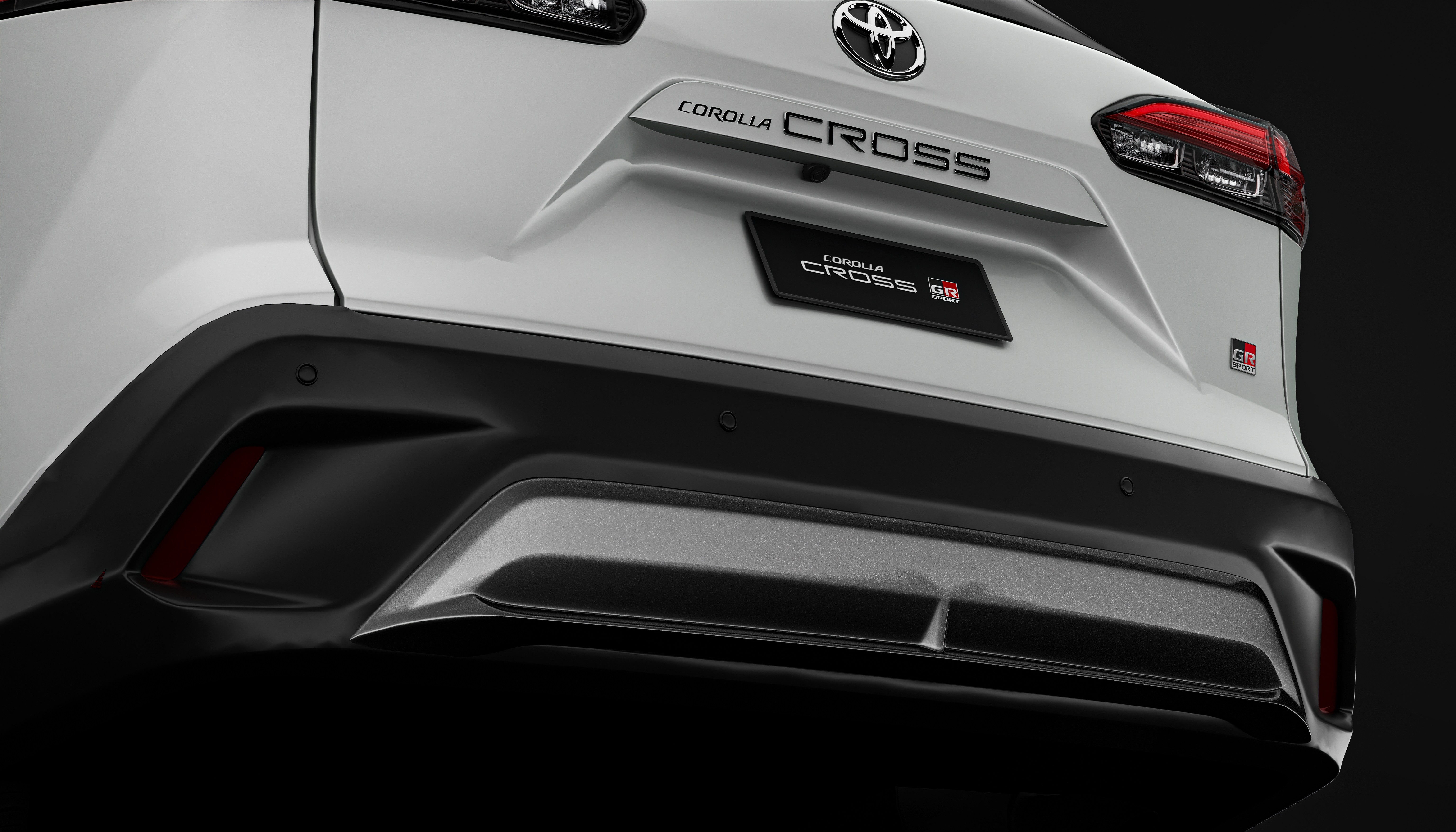 Traseira de um carro Toyota Corolla Cross branco, com para-choque preto e detalhes prateados. O logotipo da Toyota e os nomes Corolla Cross e Corolla Cross GR est&atilde;o vis&iacute;veis, junto a um emblema GR vermelho e preto. As lanternas traseiras s&atilde;o vermelhas e pretas, e h&aacute; sensores de estacionamento no para-choque. O fundo &eacute; escuro