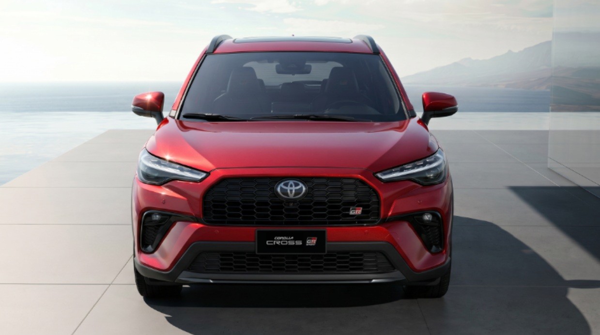 SUV Toyota Corolla Cross GR Sport vermelho, visto de frente, estacionado em um piso claro com o mar e montanhas ao fundo