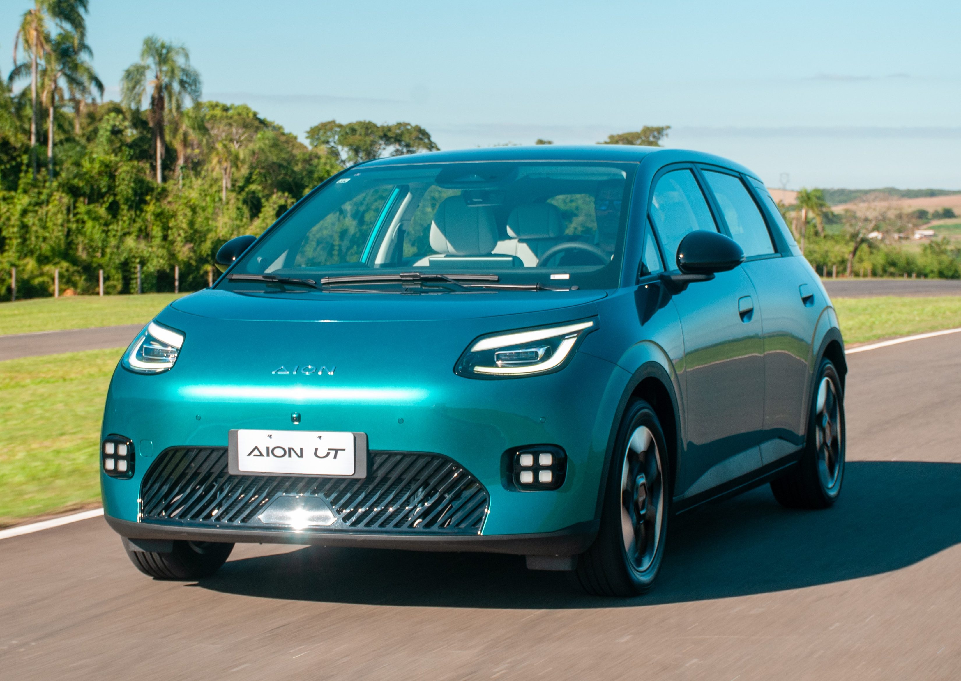 Carro el&eacute;trico Aion UT azul-turquesa em uma pista de asfalto, com vegeta&ccedil;&atilde;o exuberante e c&eacute;u azul ao fundo