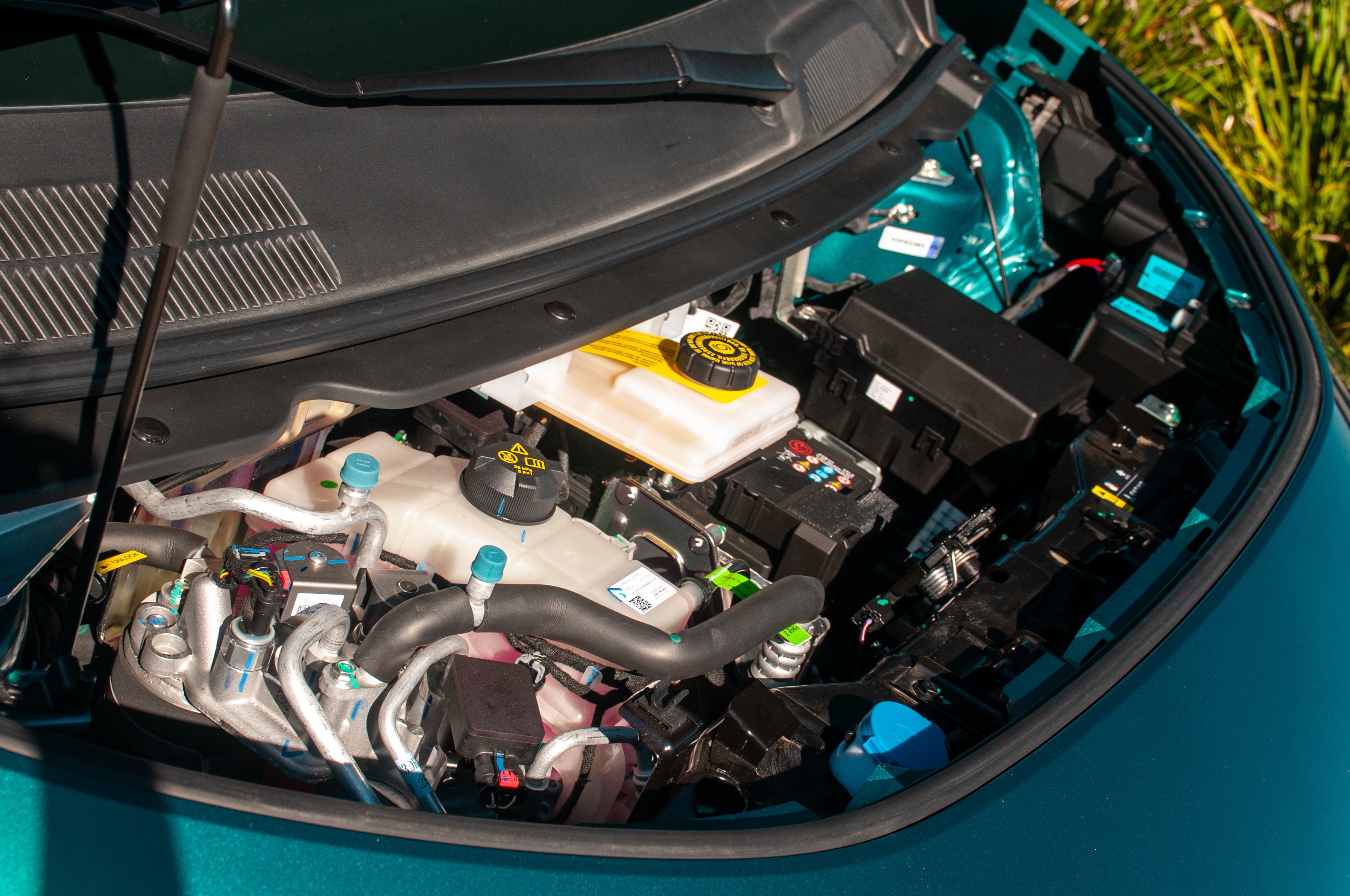Compartimento do motor de um carro, com reservat&oacute;rios de fluidos, cabos e caixas de fus&iacute;veis vis&iacute;veis, em tons de preto, branco e amarelo, sobre a lataria verde-&aacute;gua do ve&iacute;culo