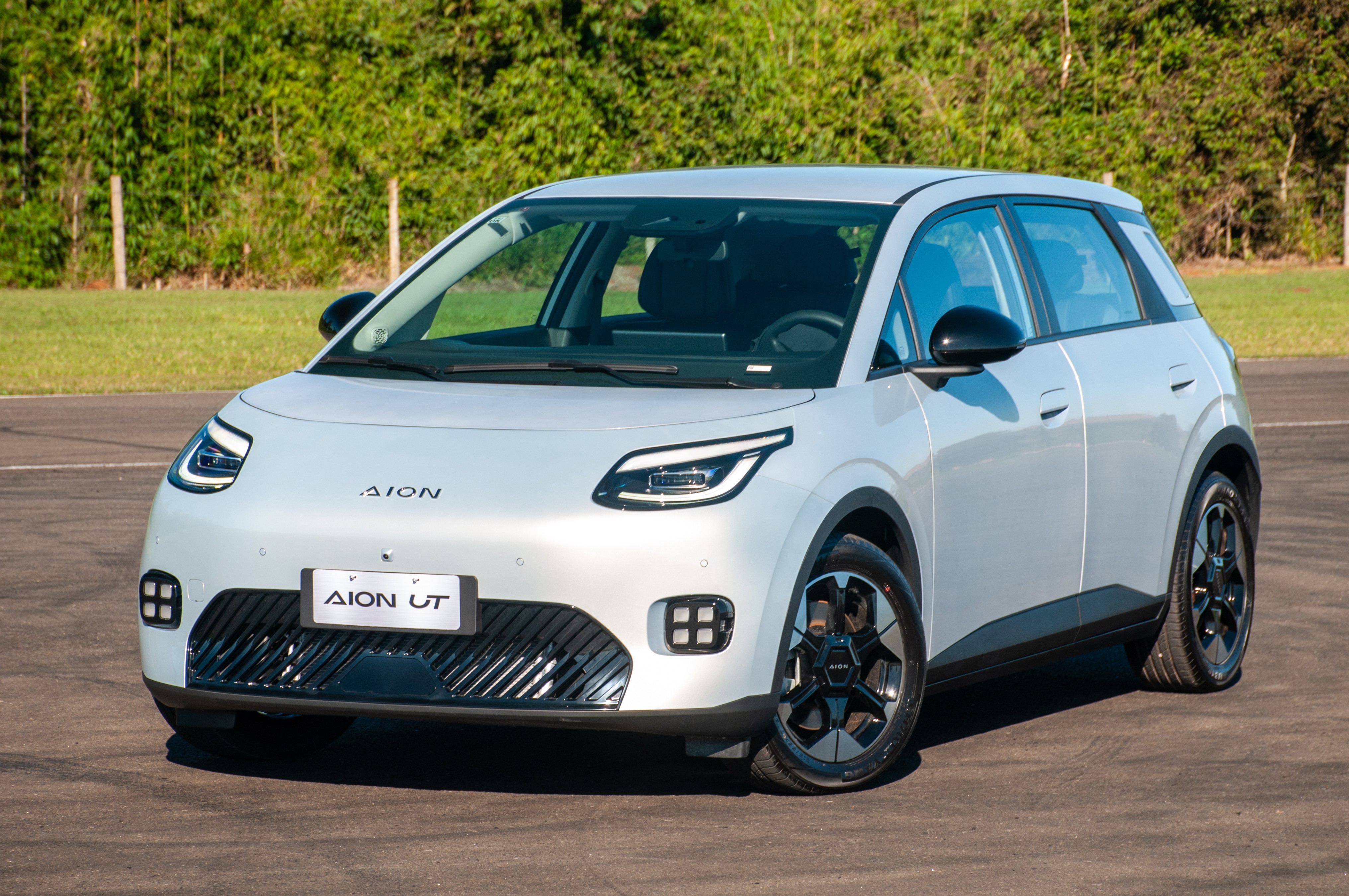 Carro el&eacute;trico Aion UT branco-prateado, com detalhes pretos nos retrovisores e rodas, estacionado em uma pista de asfalto com grama e &aacute;rvores ao fundo