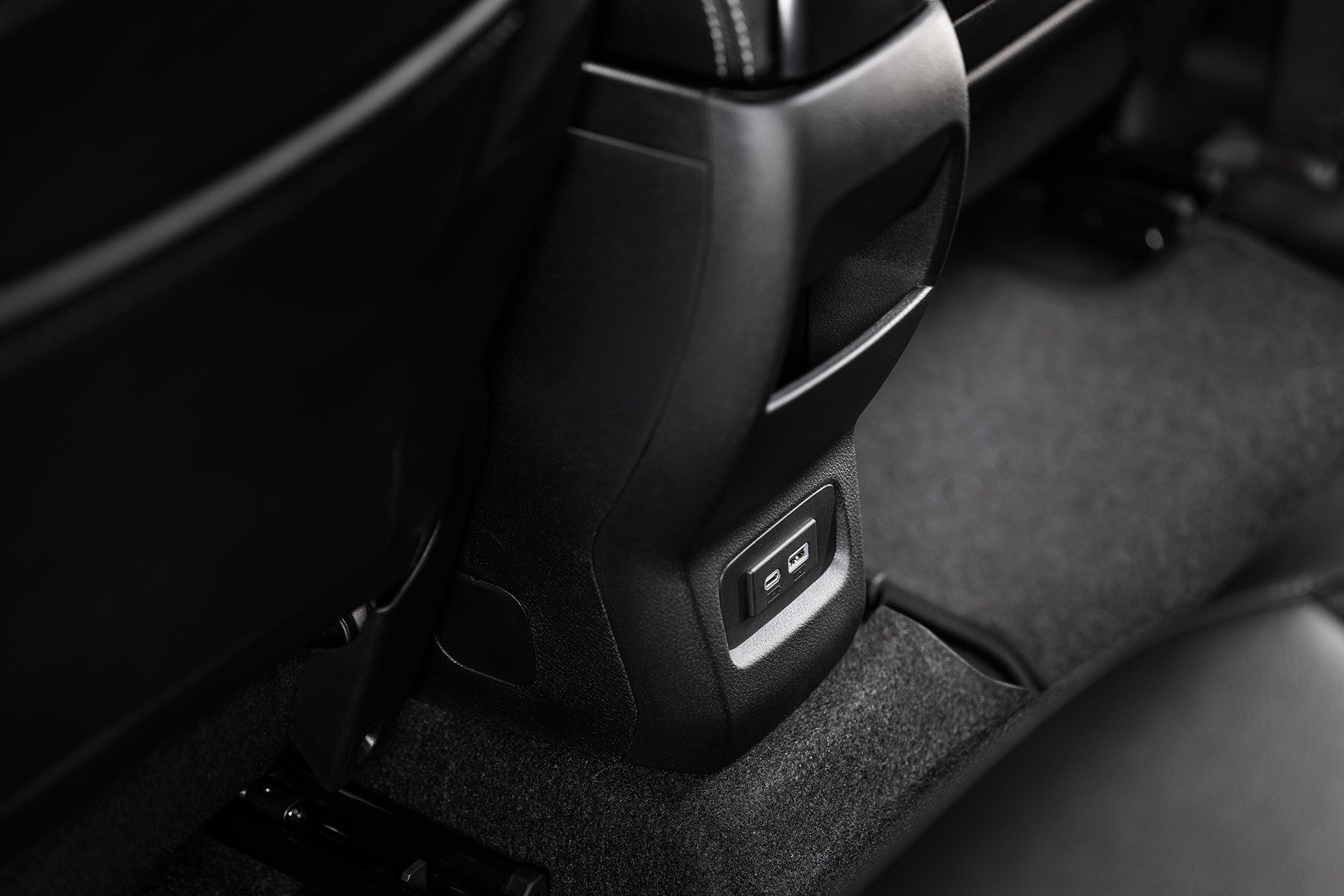 Detalhe do console central de um carro, com duas portas USB-A e USB-C para carregamento, localizadas na parte traseira, entre os bancos dianteiros. O interior &eacute; predominantemente preto, com estofamento e carpete escuros
