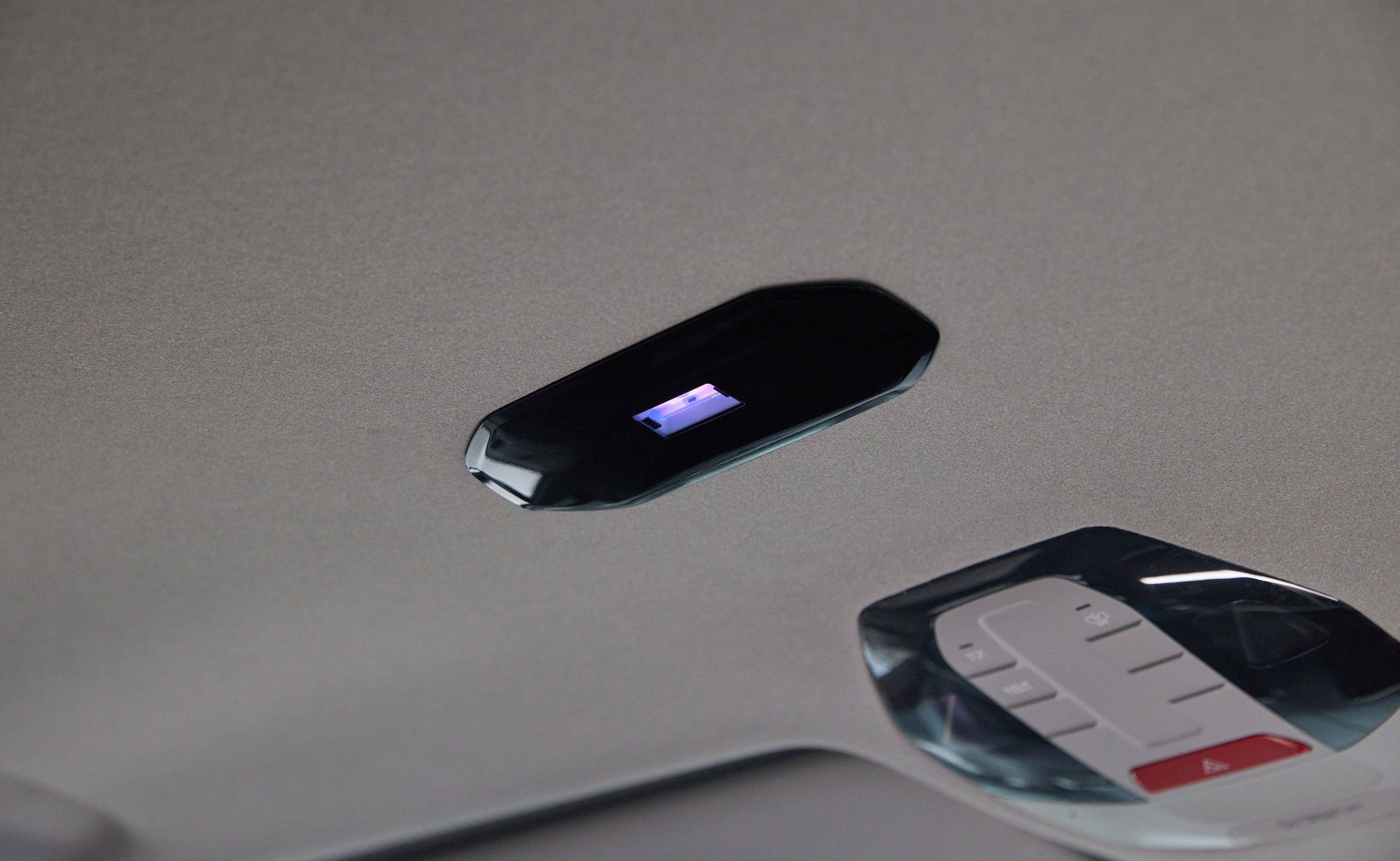 Painel de controle no teto de um carro, com uma tela retangular preta exibindo luz azul e um conjunto de bot&otilde;es brancos e um vermelho