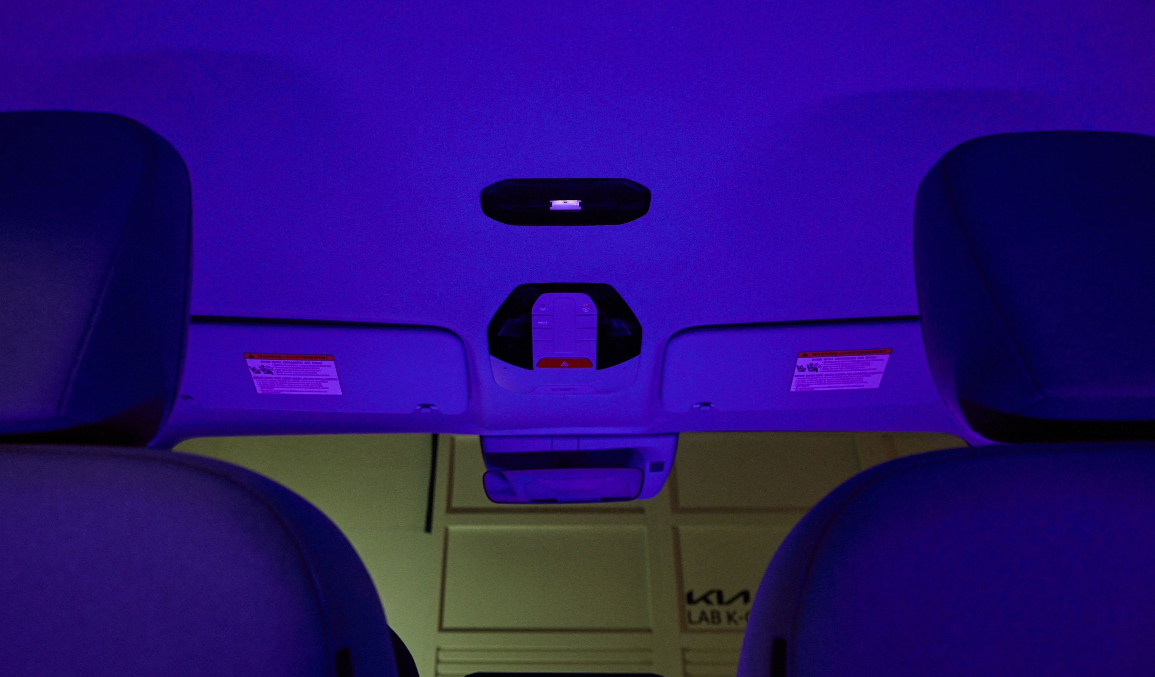 Interior de um carro com ilumina&ccedil;&atilde;o ambiente roxa, vista de tr&aacute;s dos bancos dianteiros. O teto e os para-s&oacute;is est&atilde;o vis&iacute;veis, com uma luz de teto central e um espelho retrovisor. Ao fundo, uma parede clara com o logo KIA LAB K aparece