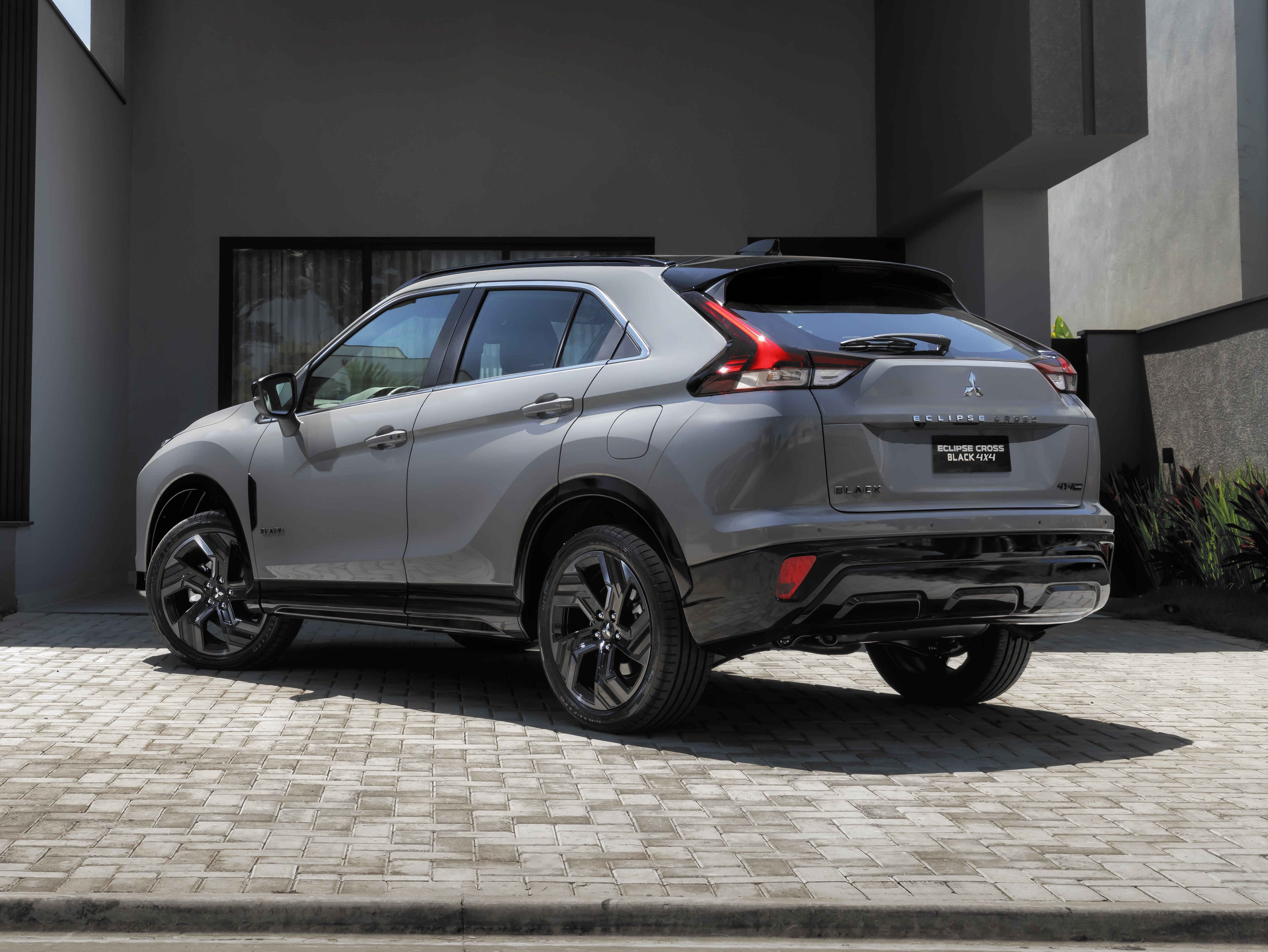 SUV Mitsubishi Eclipse Cross cinza-claro estacionado de r&eacute; na entrada de uma casa moderna com garagem coberta. O carro tem rodas pretas e detalhes escuros. &Agrave; esquerda, um coqueiro e gramado verde. A casa tem fachada em tons de cinza e preto, com grandes janelas e design angular. O c&eacute;u est&aacute; azul e claro