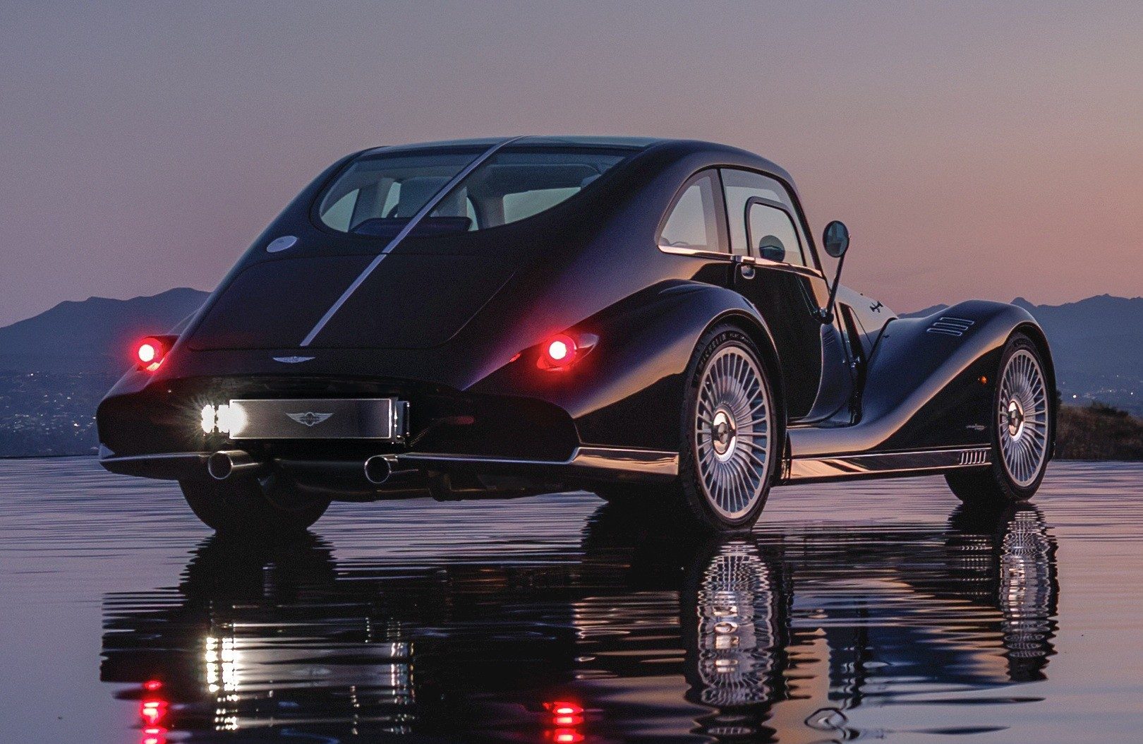 Carro esportivo Morgan Plus Four preto, com design retr&ocirc;, visto por tr&aacute;s e ligeiramente de lado, parado em uma superf&iacute;cie molhada que reflete suas luzes traseiras vermelhas e a placa iluminada. Ao fundo, montanhas escuras sob um c&eacute;u crepuscular em tons de roxo e laranja
