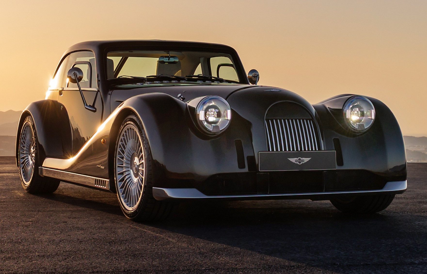 Carro esportivo Morgan Plus Four preto brilhante, com far&oacute;is acesos e rodas prateadas, estacionado em uma estrada escura ao p&ocirc;r do sol, com montanhas ao fundo