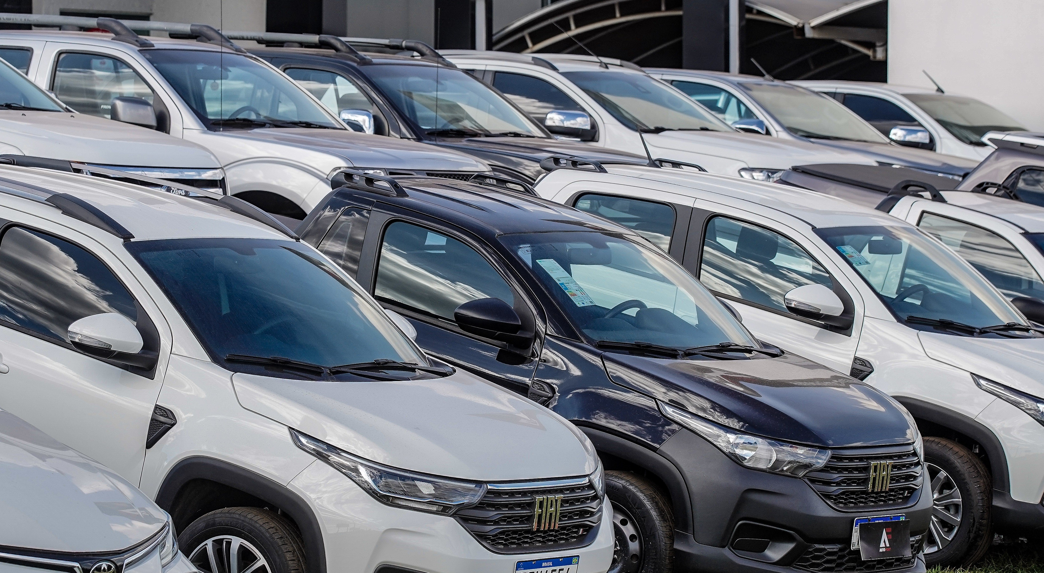 Fileira de carros novos, incluindo SUVs e picapes, estacionados ao ar livre. H&aacute; ve&iacute;culos brancos e um preto em destaque no centro, com detalhes em cinza escuro e rack de teto. O c&eacute;u reflete nos para-brisas, sugerindo um dia claro.