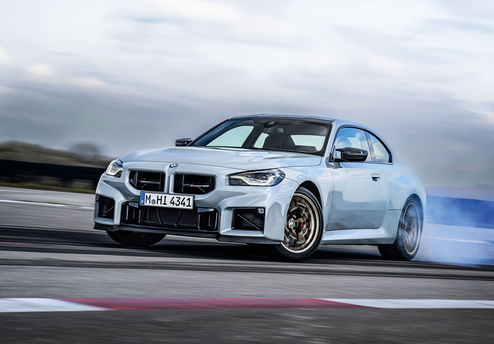 Carro esportivo BMW M2 cinza-claro em movimento, derrapando em uma pista de corrida, com fumaça azul saindo do pneu traseiro direito. O céu está nublado