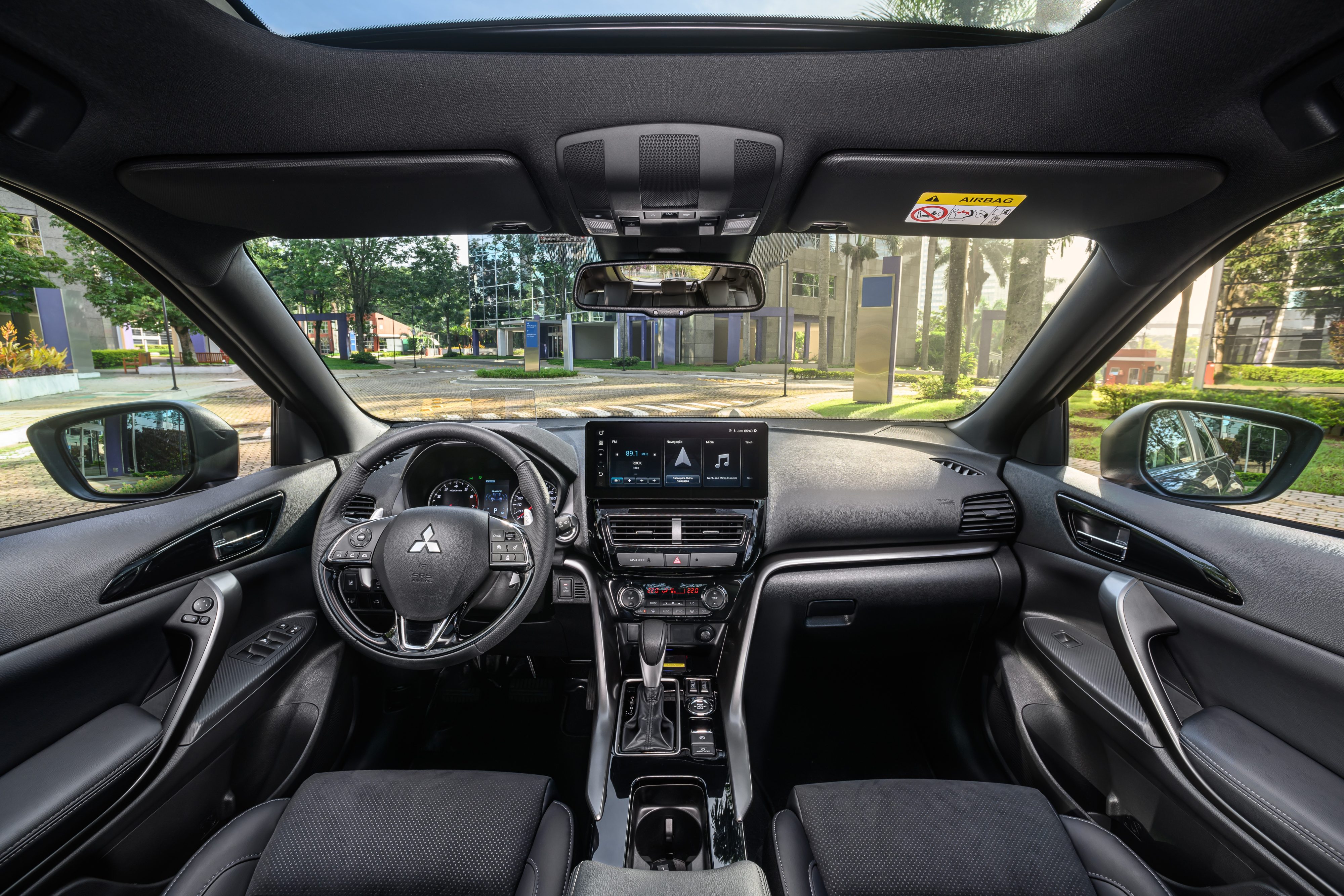 Interior de um carro moderno com bancos pretos, volante multifuncional e painel com tela multim&iacute;dia. O para-brisa revela uma rua arborizada com pr&eacute;dios e postes sob luz solar