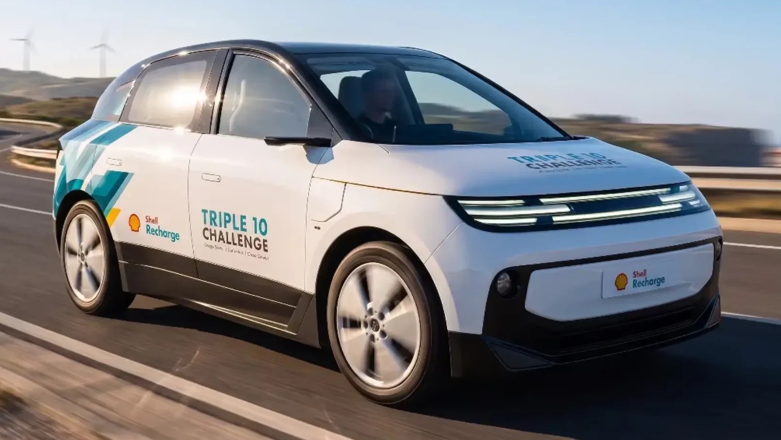 Carro elétrico branco com teto preto e detalhes azuis e verdes, logotipos Shell Recharge e Triple 10 Challenge, em movimento numa estrada com turbinas eólicas ao fundo