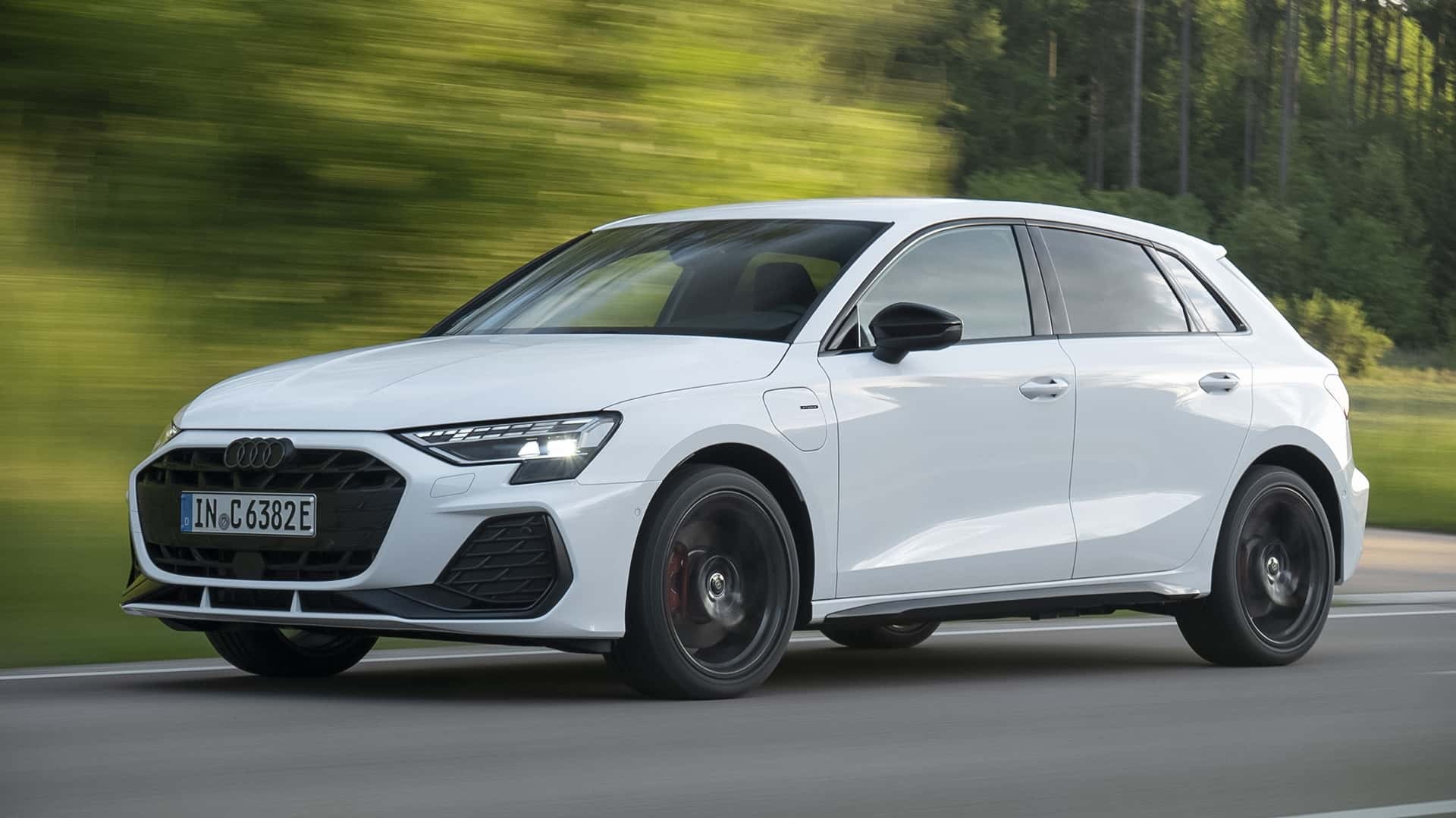 Carro Audi A3 branco em movimento, visto de lado, com far&oacute;is acesos, em uma estrada com &aacute;rvores verdes borradas ao fundo