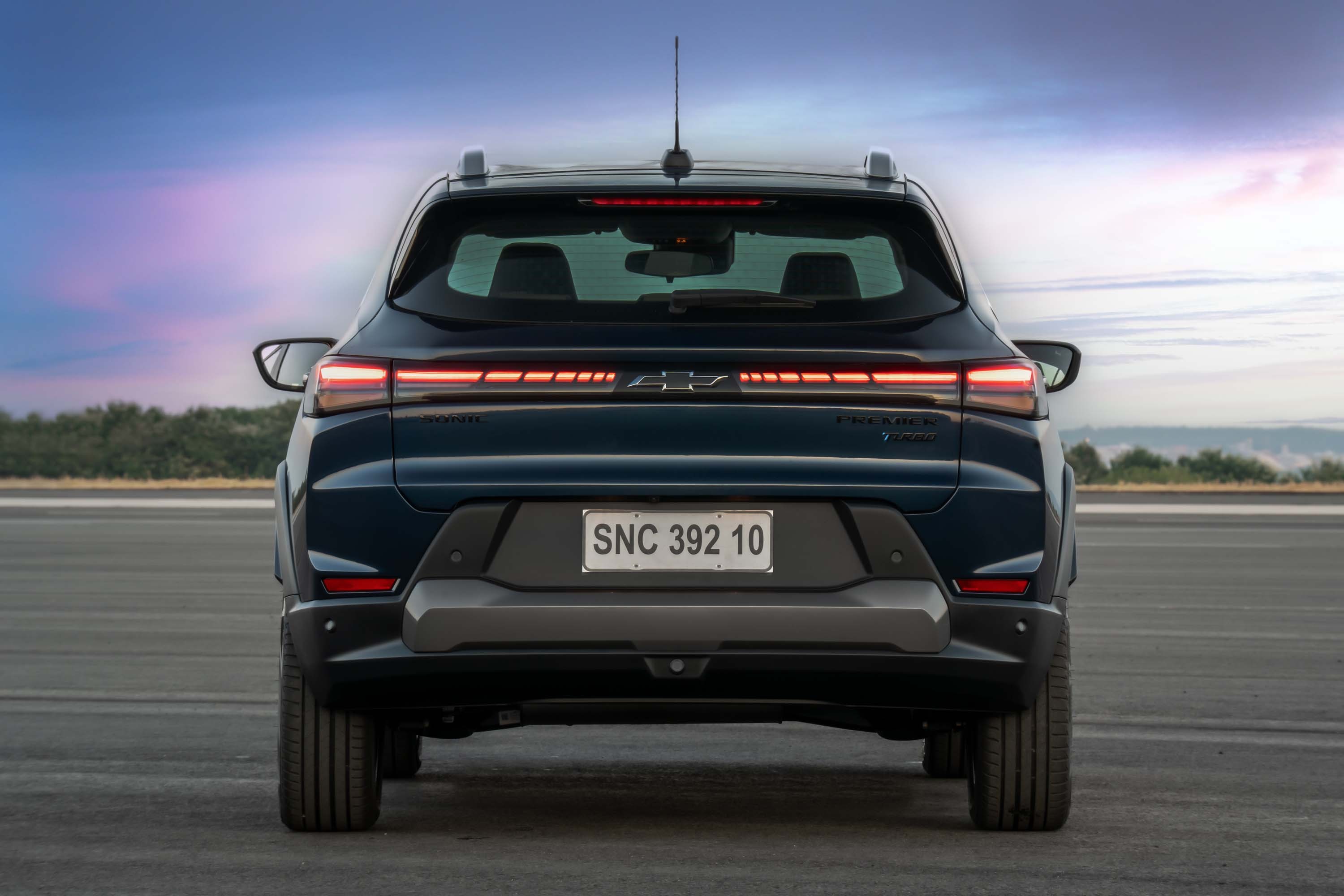 Traseira de um carro SUV azul-escuro, com lanternas horizontais finas e acesas, sobre as quais est&aacute; o emblema Chevrolet. A placa branca exibe SNC 392 10. O para-choque traseiro &eacute; cinza-escuro, e o ve&iacute;culo est&aacute; em uma pista de asfalto, com um c&eacute;u azul e nuvens ao fundo