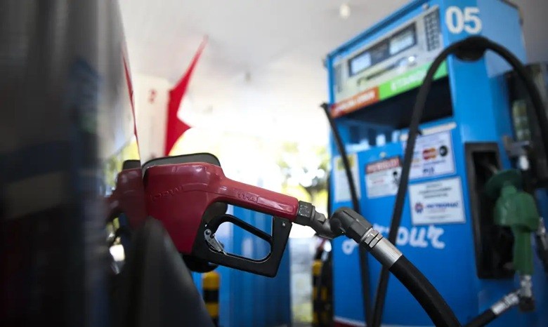 Bico de combust&iacute;vel vermelho inserido no tanque de um ve&iacute;culo, com uma bomba de gasolina azul e branca ao fundo, em um posto de combust&iacute;vel