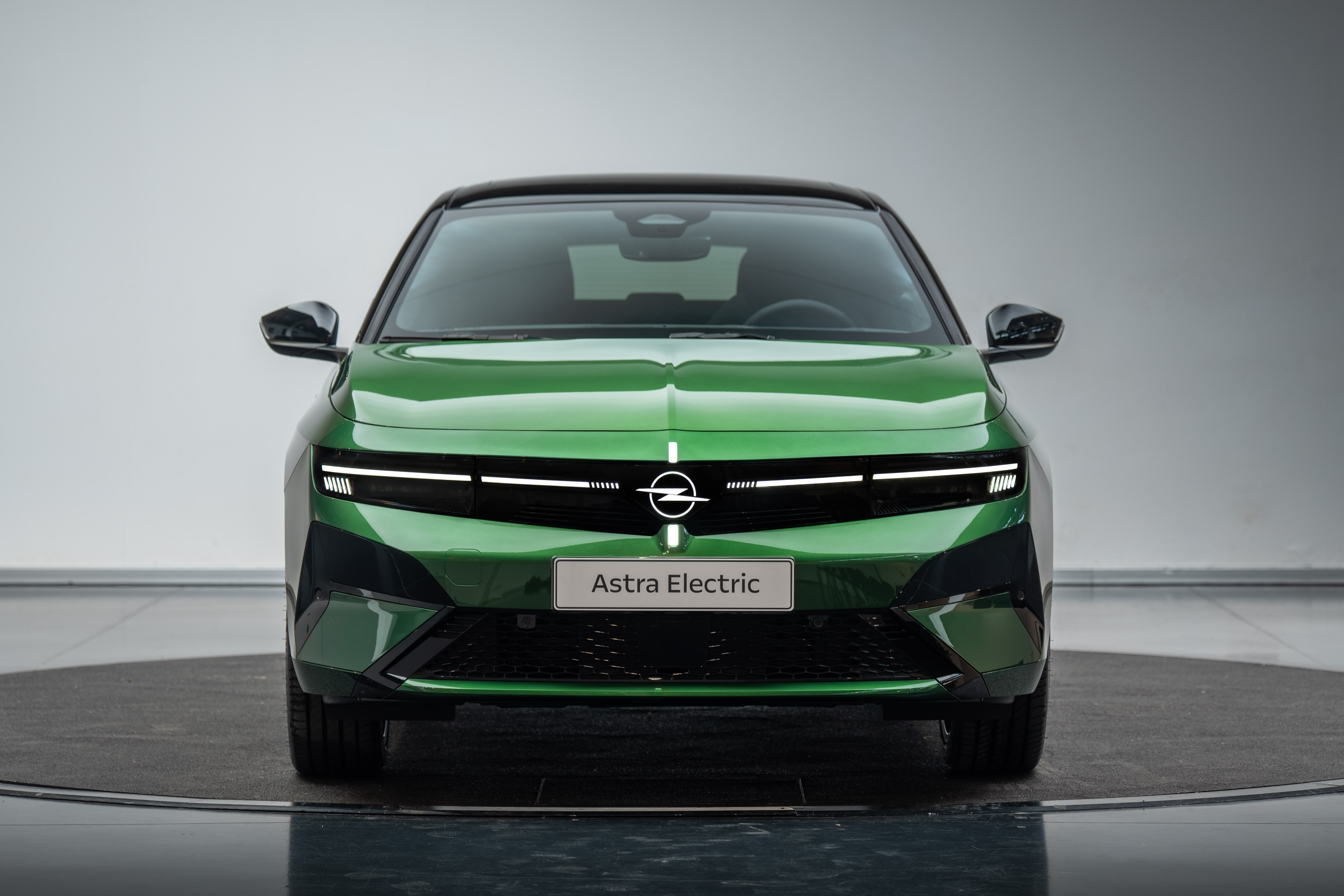 Carro Opel Astra Electric verde met&aacute;lico, visto de frente, com teto preto e far&oacute;is de LED horizontais acesos, exibindo a placa Astra Electric no para-choque, sobre uma plataforma girat&oacute;ria cinza em um est&uacute;dio branco